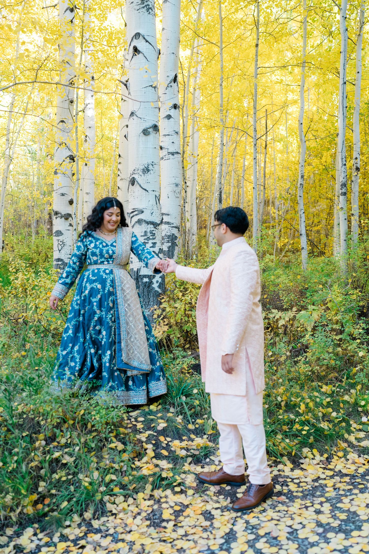 Two people walk among golden aspen trees in autumn with fallen yellow leaves scattered on the ground.