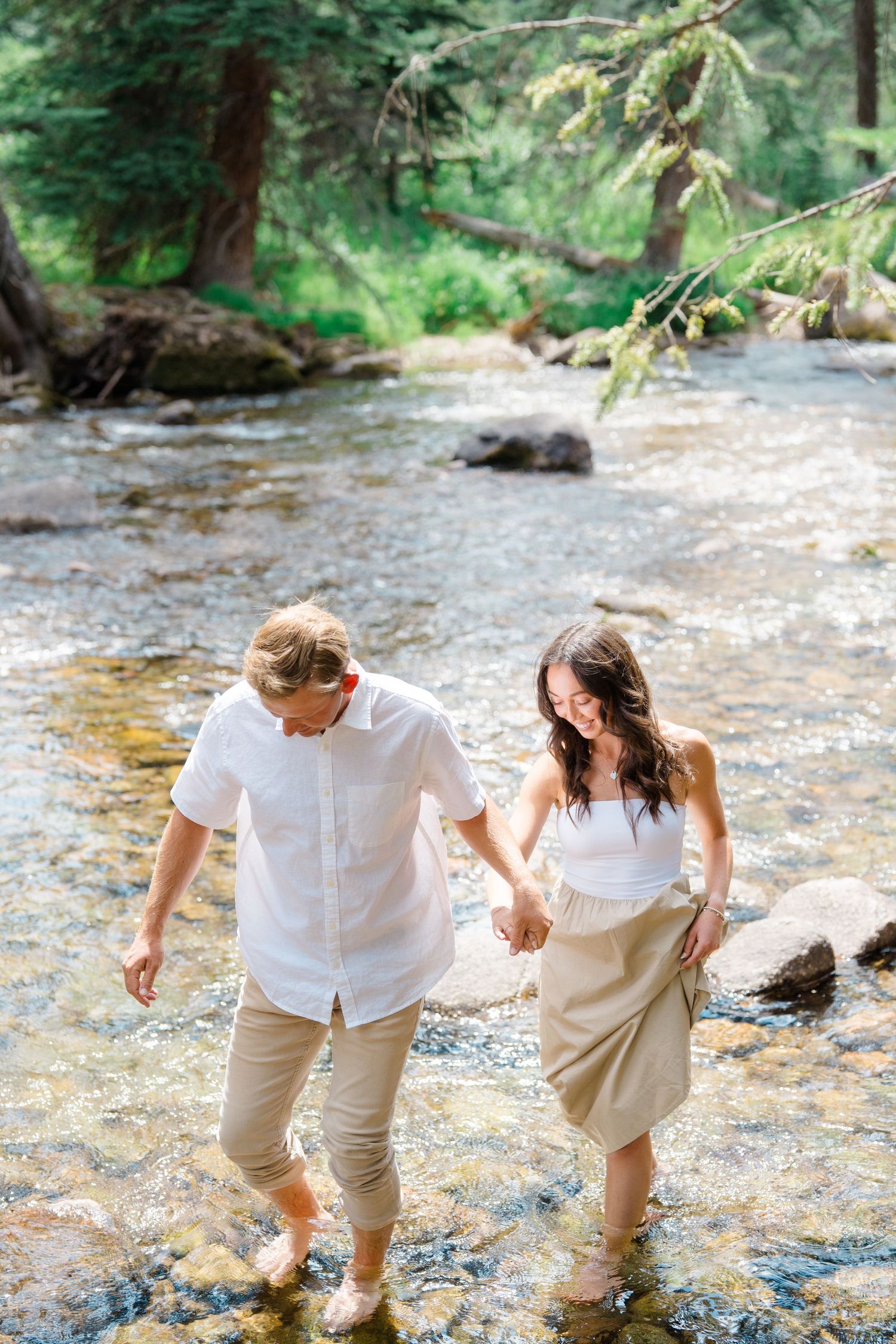 A playful couple splashes and laughs while walking through a shallow creek, dressed in matching white and khaki outfits.