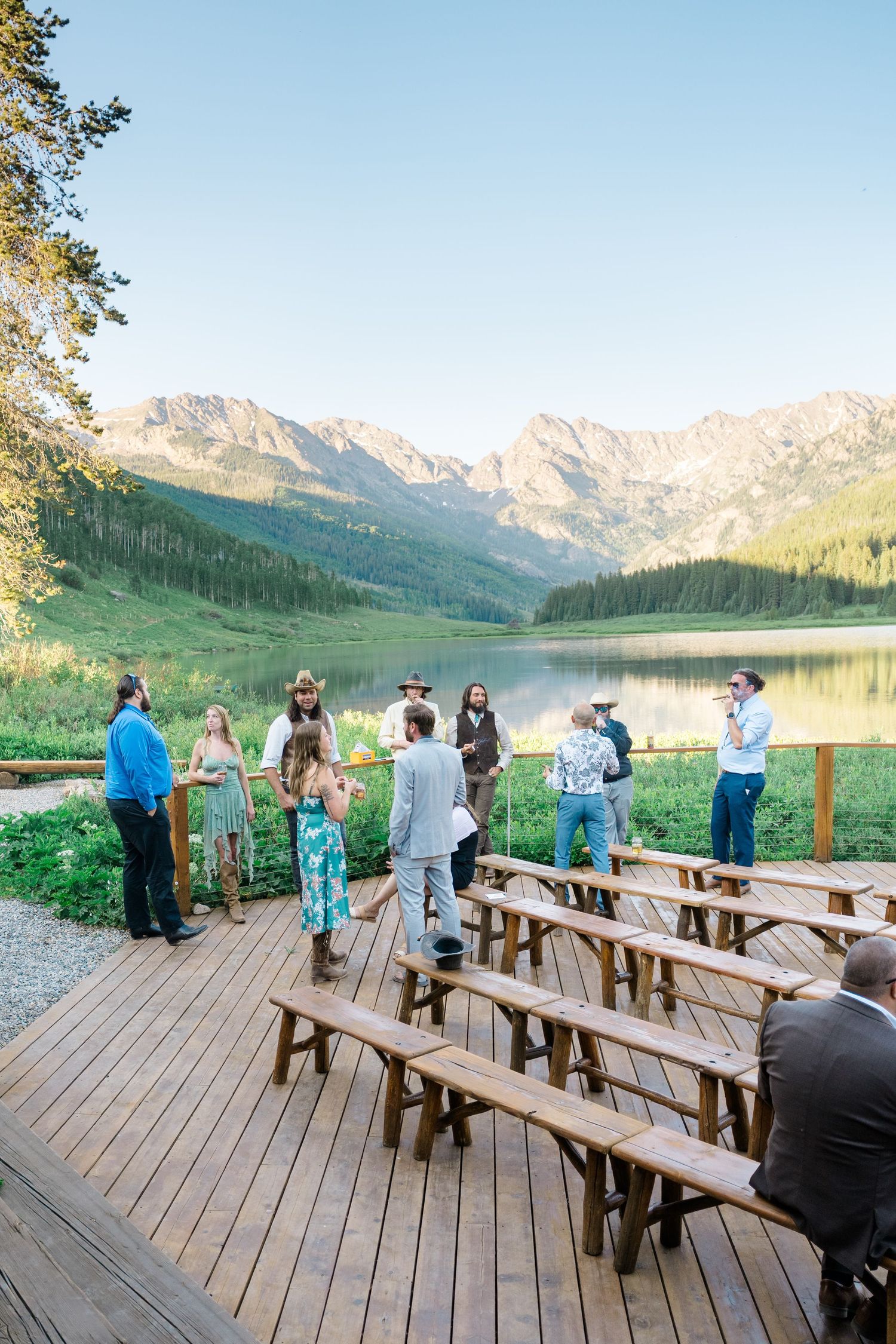 Wedding guests mingling on a wooden deck overlooking a mountain lake during golden hour at Piney River Ranch Wedding.