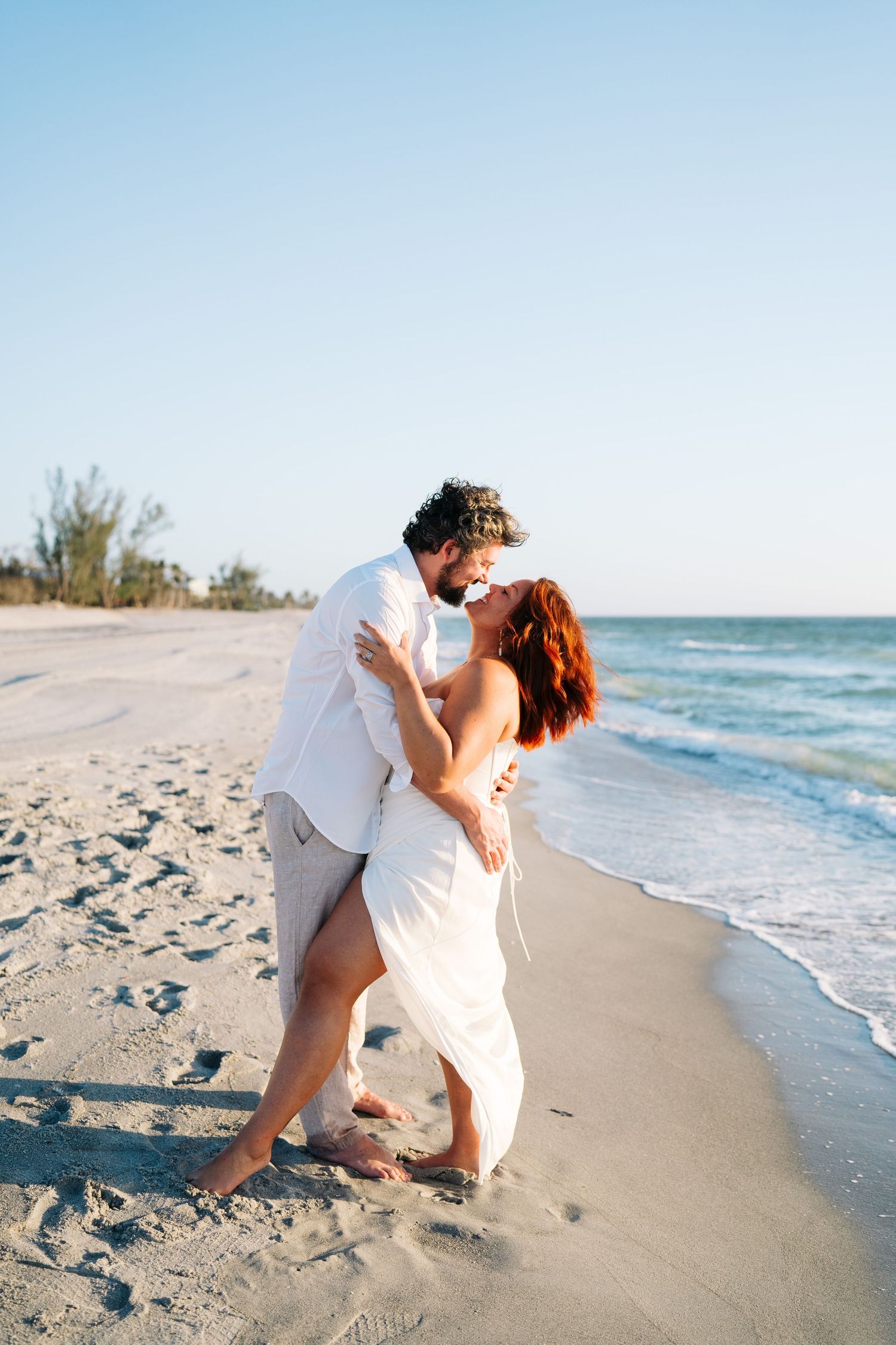 Playful couple in white outfits dancing and laughing together on a sandy beach at sunset.