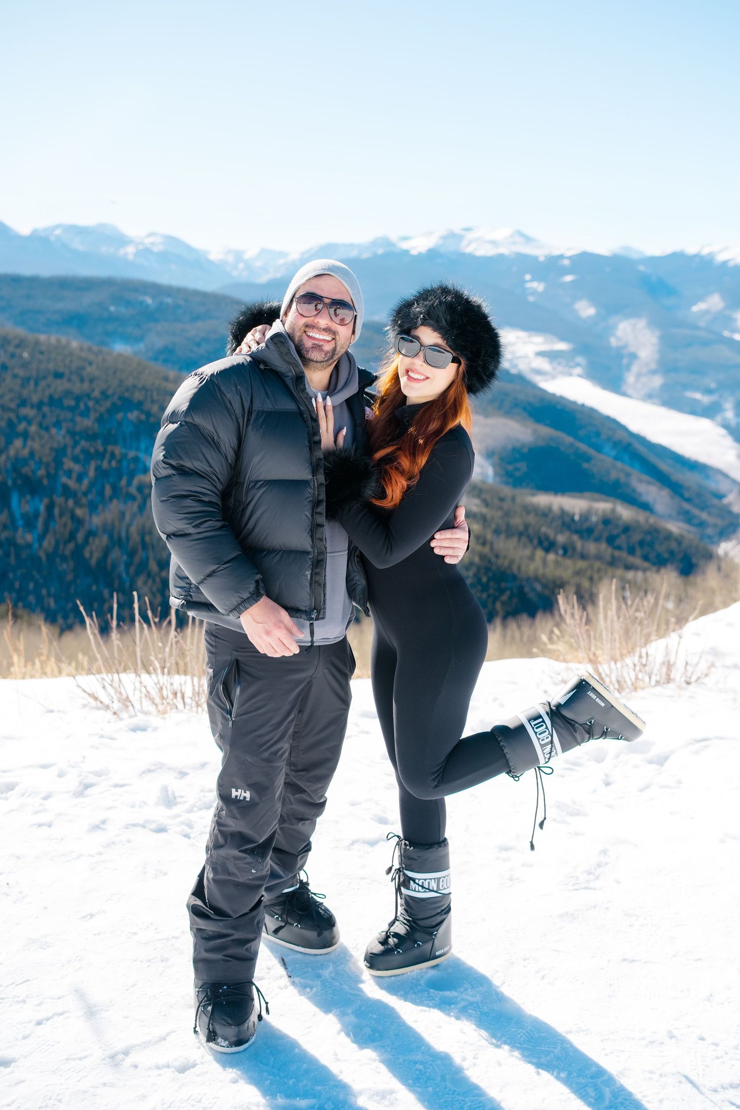 A couple in winter gear poses playfully in the snow with snow-capped mountains and forest in the background.
