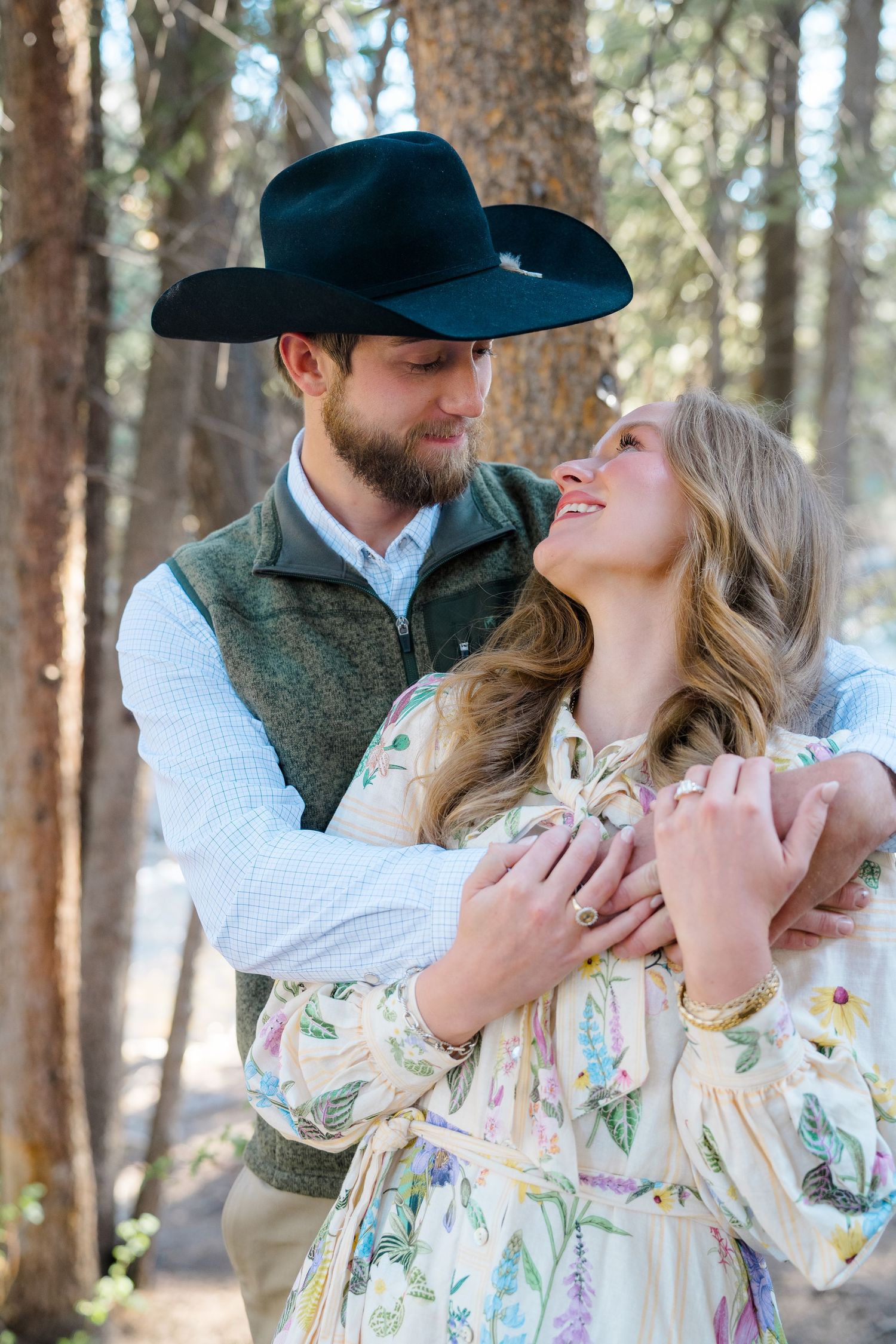 A couple embraces in a pine forest, man in black cowboy hat and vest holds smiling blonde woman in floral dress.