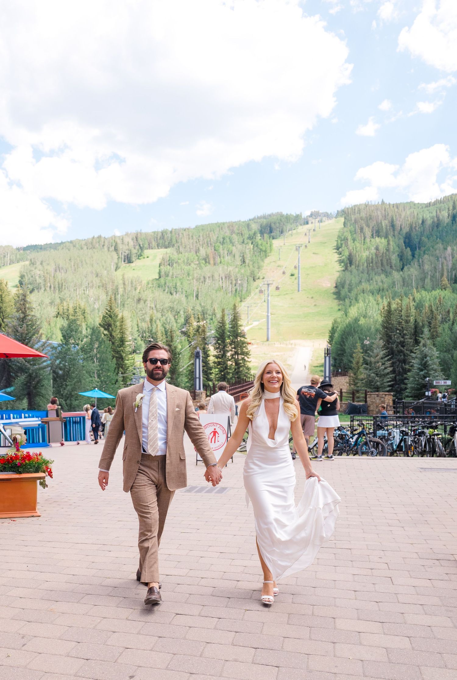 A bride and groom walk hand-in-hand at a mountain ski resort wedding venue in summer, surrounded by lush green slopes.
