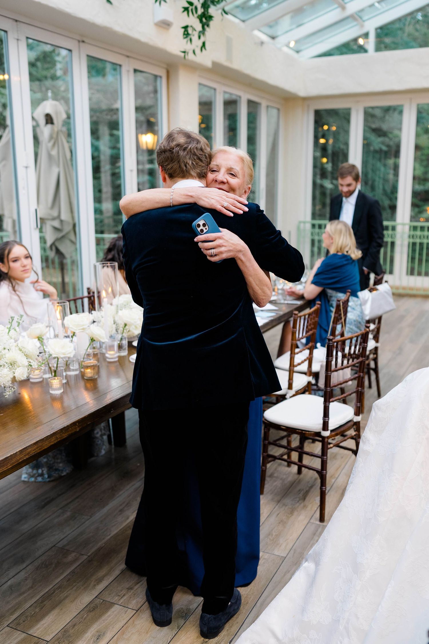 Two guests share a warm embrace at an elegant wedding reception in a glass conservatory with floral centerpieces.