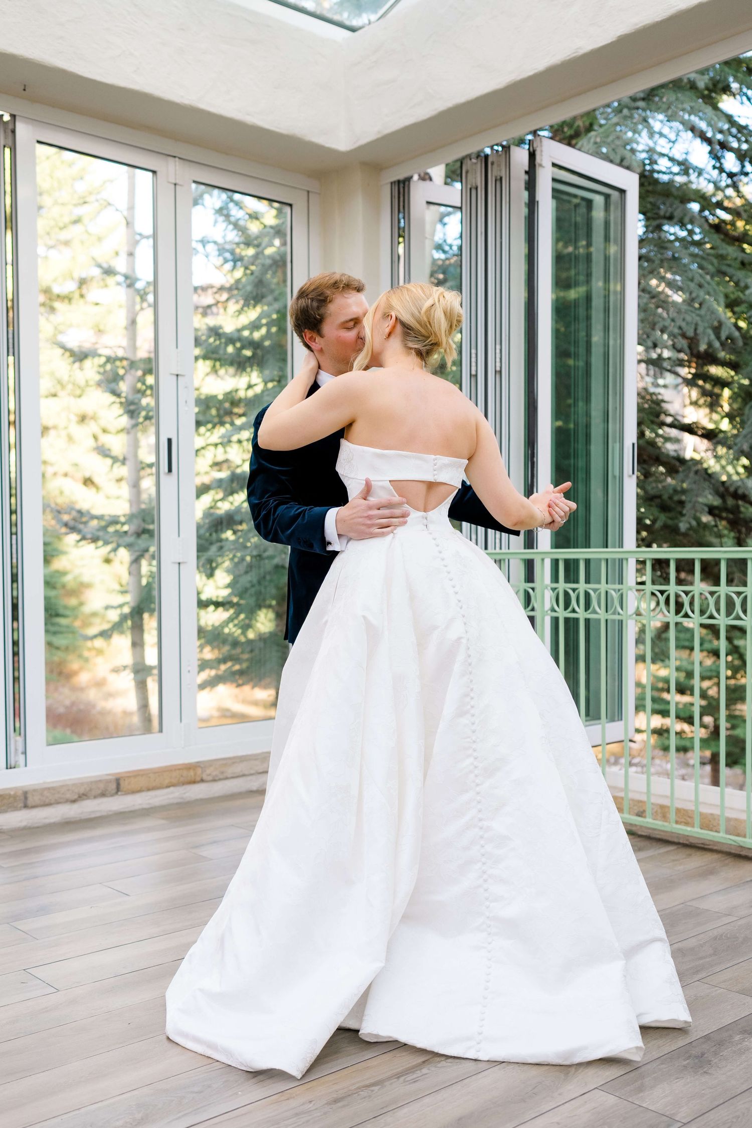 Bride and groom share a romantic first dance in a sunlit room with large windows and lush green trees outside.
