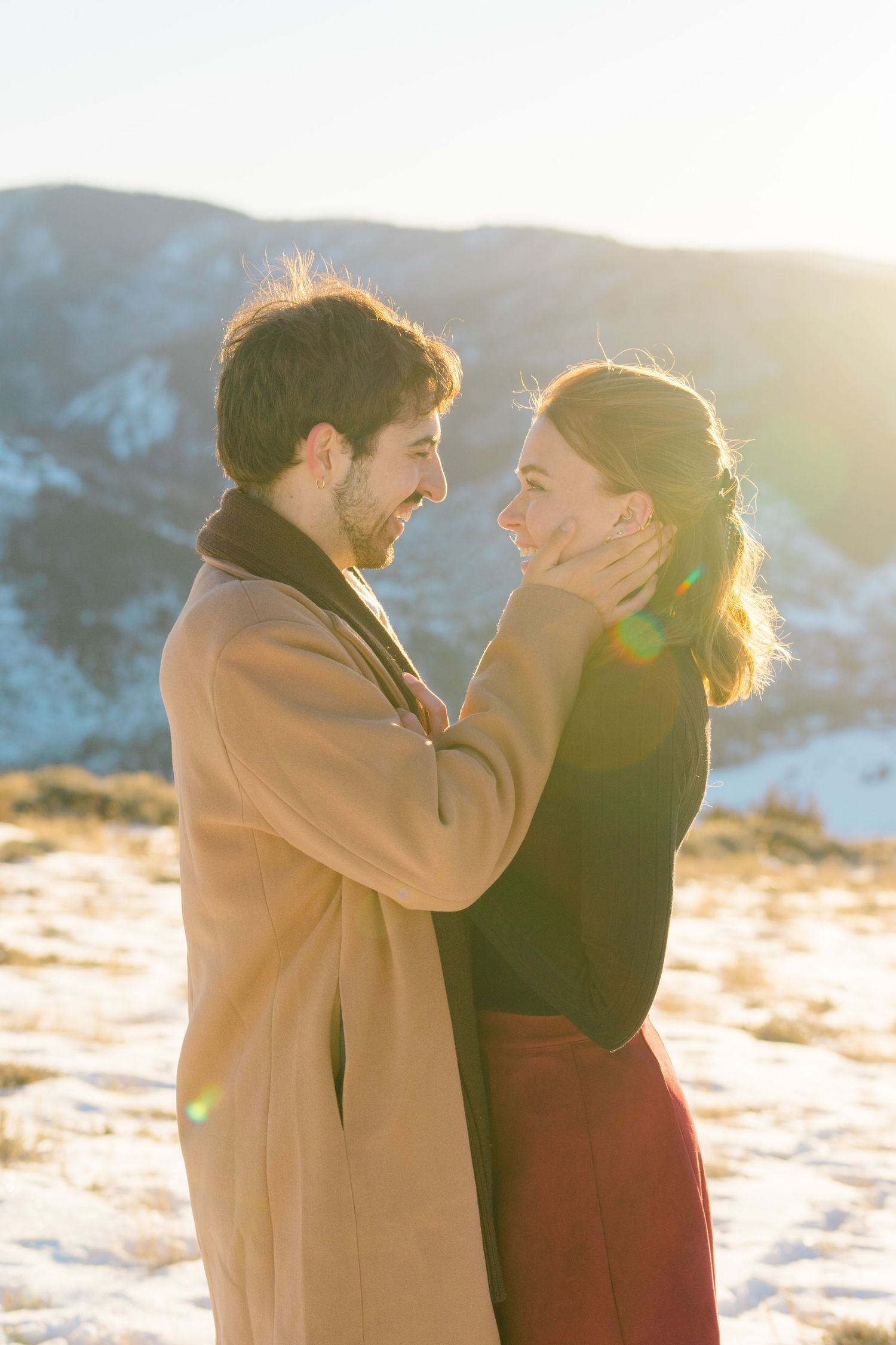 A couple shares a tender moment in the winter sunlight with snow on the ground and mountains in the background.