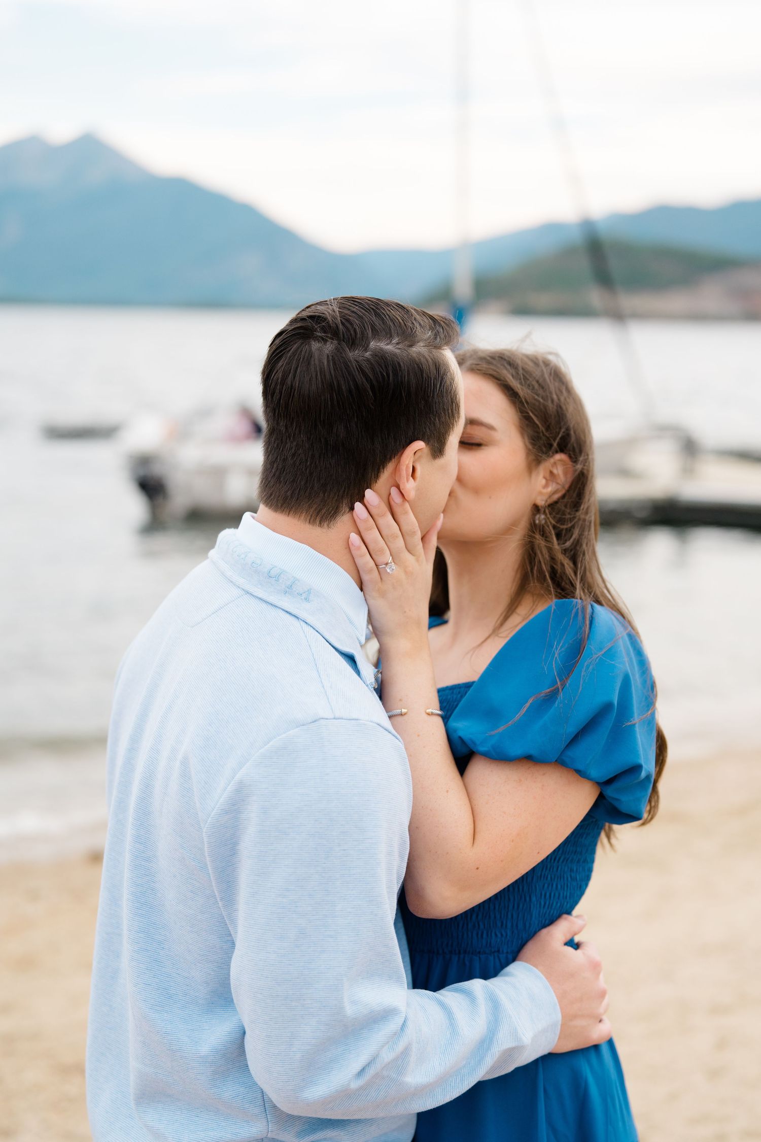 A romantic moment captured at a lakeside beach with mountains in the background.