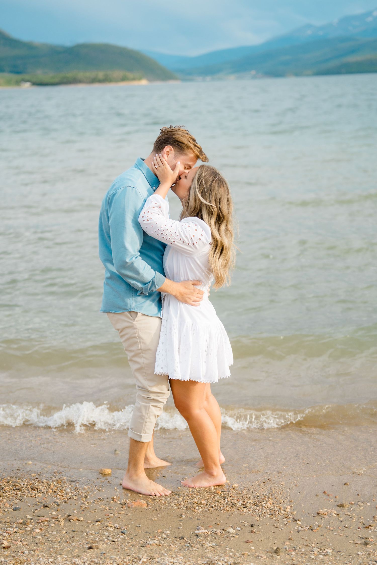 A romantic couple embraces at the water's edge on a beach with mountains in the background.