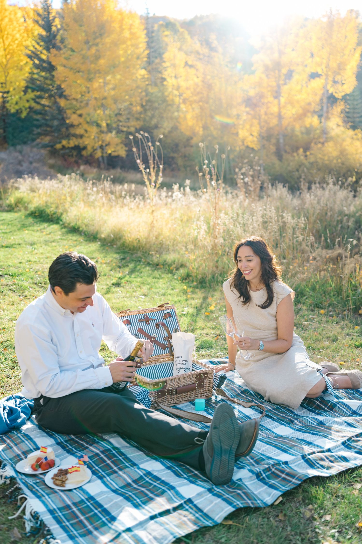 A couple enjoys a romantic fall picnic on a blue plaid blanket surrounded by golden autumn foliage.
