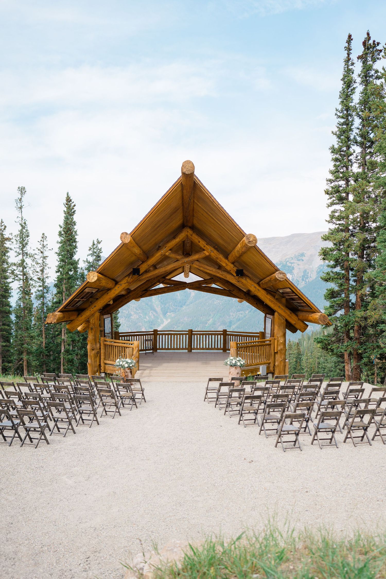 Outdoor mountain wedding ceremony setup with wooden arch and folding chairs against a backdrop of evergreen trees and peaks.