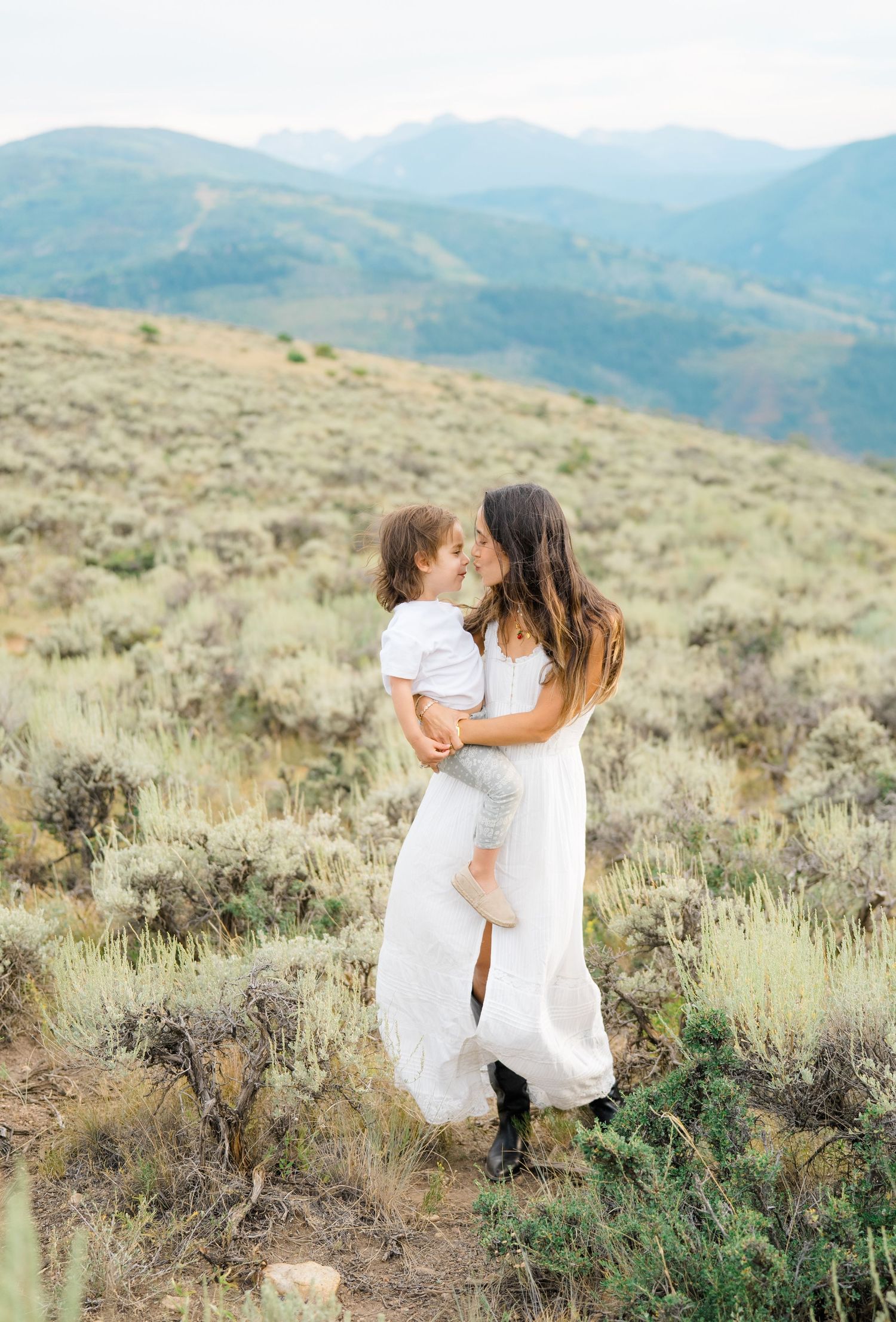 Two figures in white dresses dance and embrace among desert sagebrush with mountains in background.