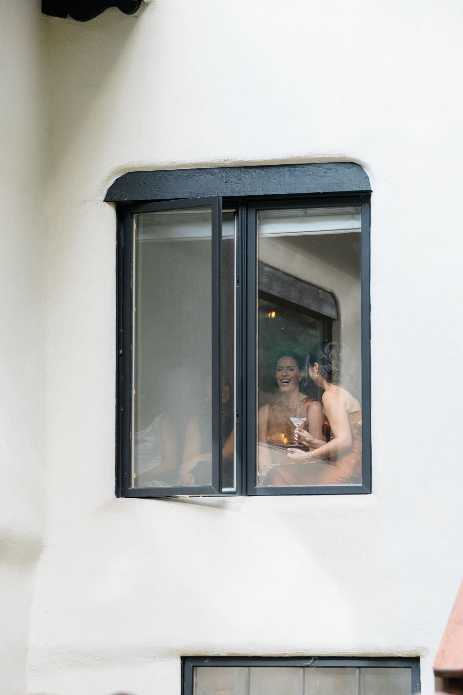 A view through a window shows a couple sharing an intimate moment in a white room.