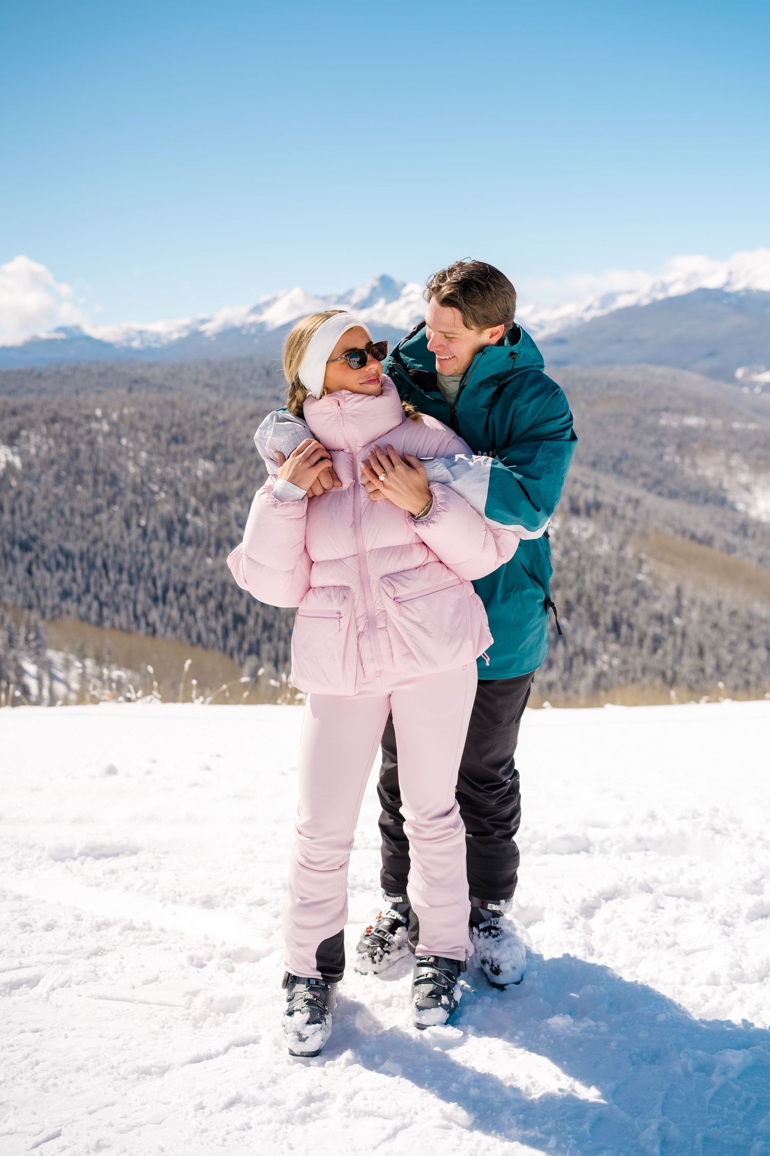 A couple in winter attire shares playful moments in the snow with mountains visible in the background.