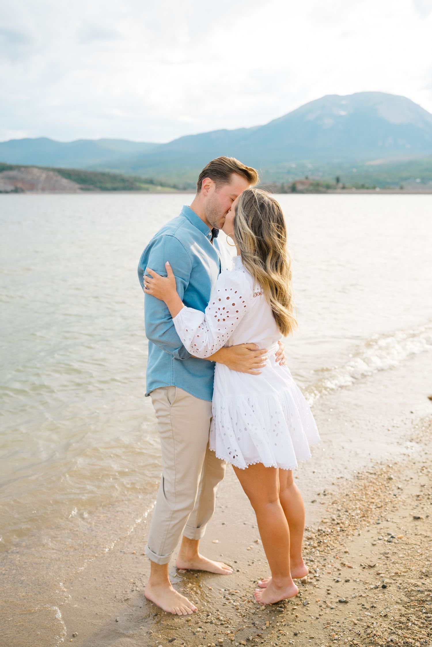 A couple embraces and kisses on a scenic beach with mountains in the background during vail proposal photography session.