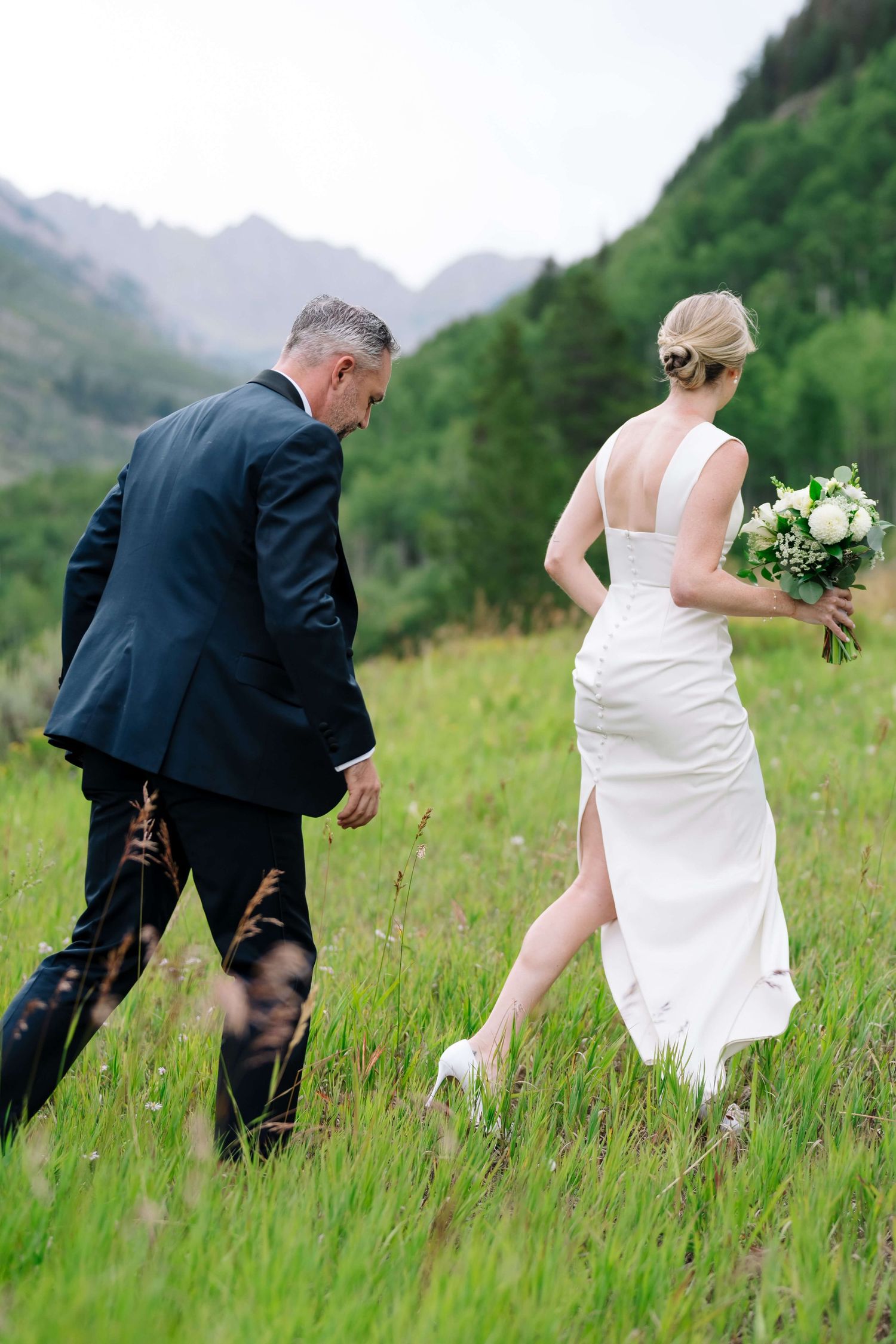 Bride and groom walking through green mountain meadow at Vail Golf Course - bride holding white bouquet in elegant backless wedding dress.