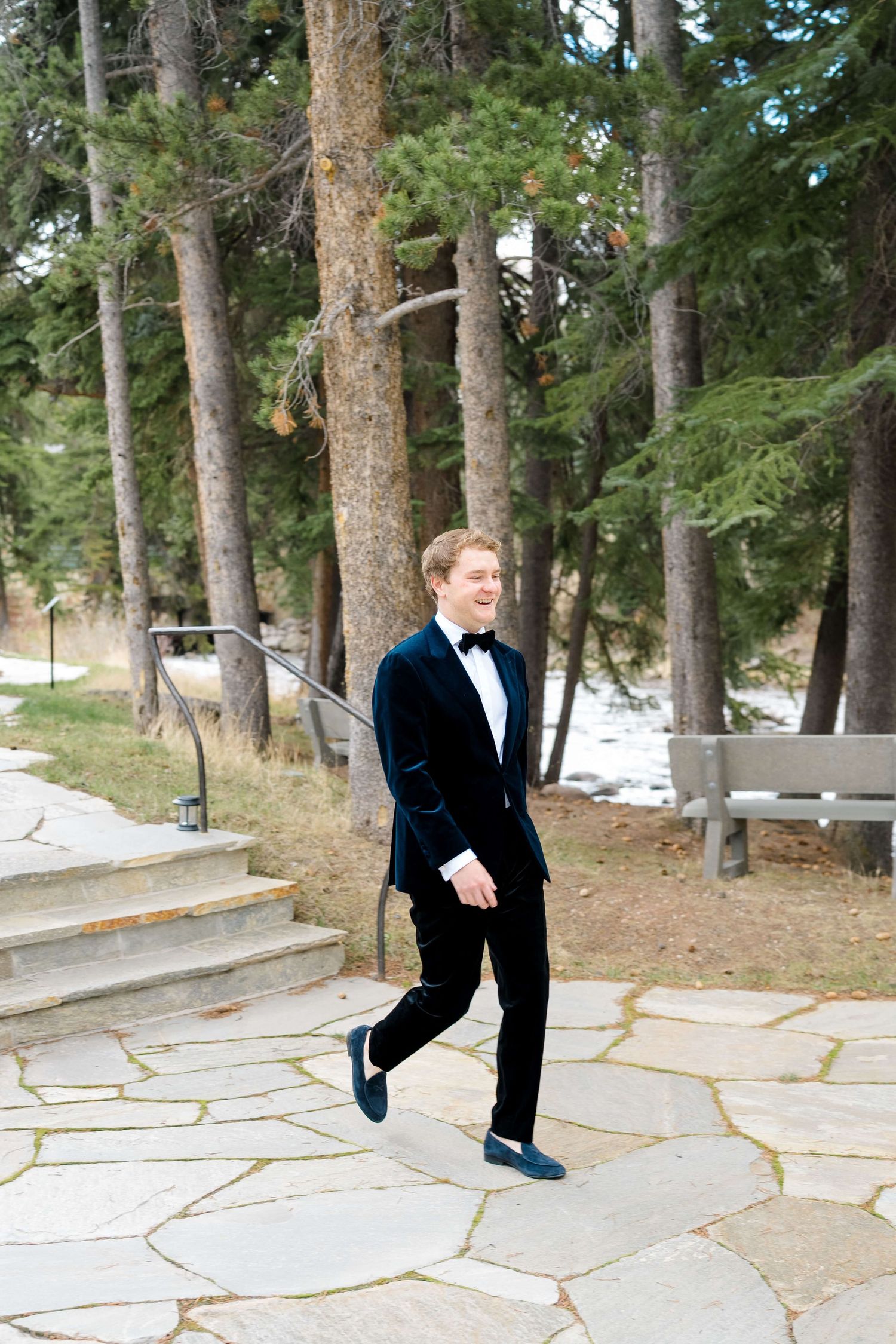 Groom in navy blue velvet tuxedo and bow tie walking on stone path outdoors among pine trees.