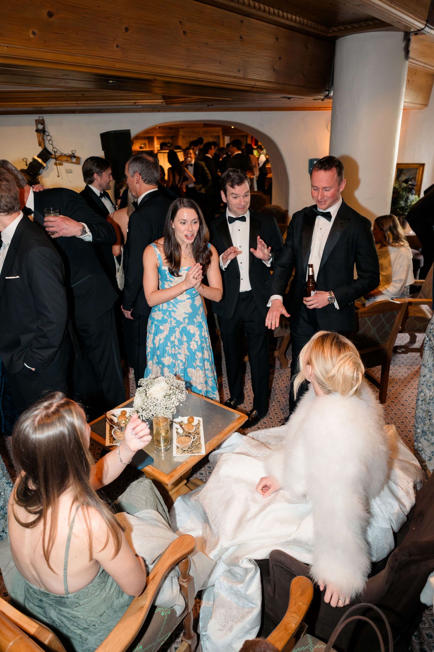 Elegant formal event with guests in tuxedos and gowns socializing around a table in a warmly lit venue.