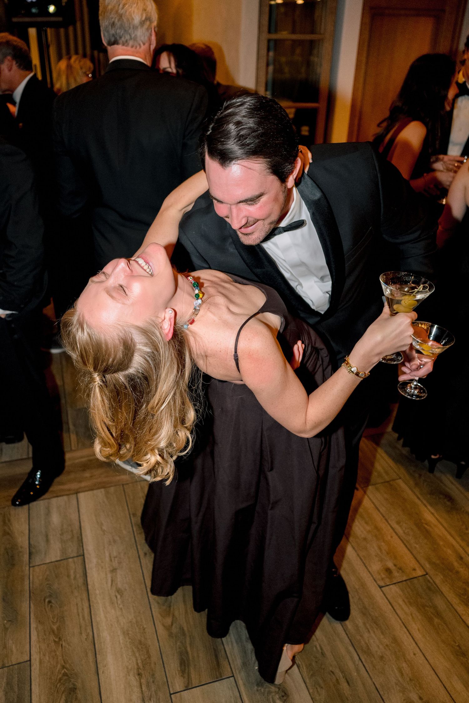Couple dancing and laughing at a formal event, man in tuxedo holding champagne glass, woman in elegant dark gown at Vail Sonnenalp Wedding.