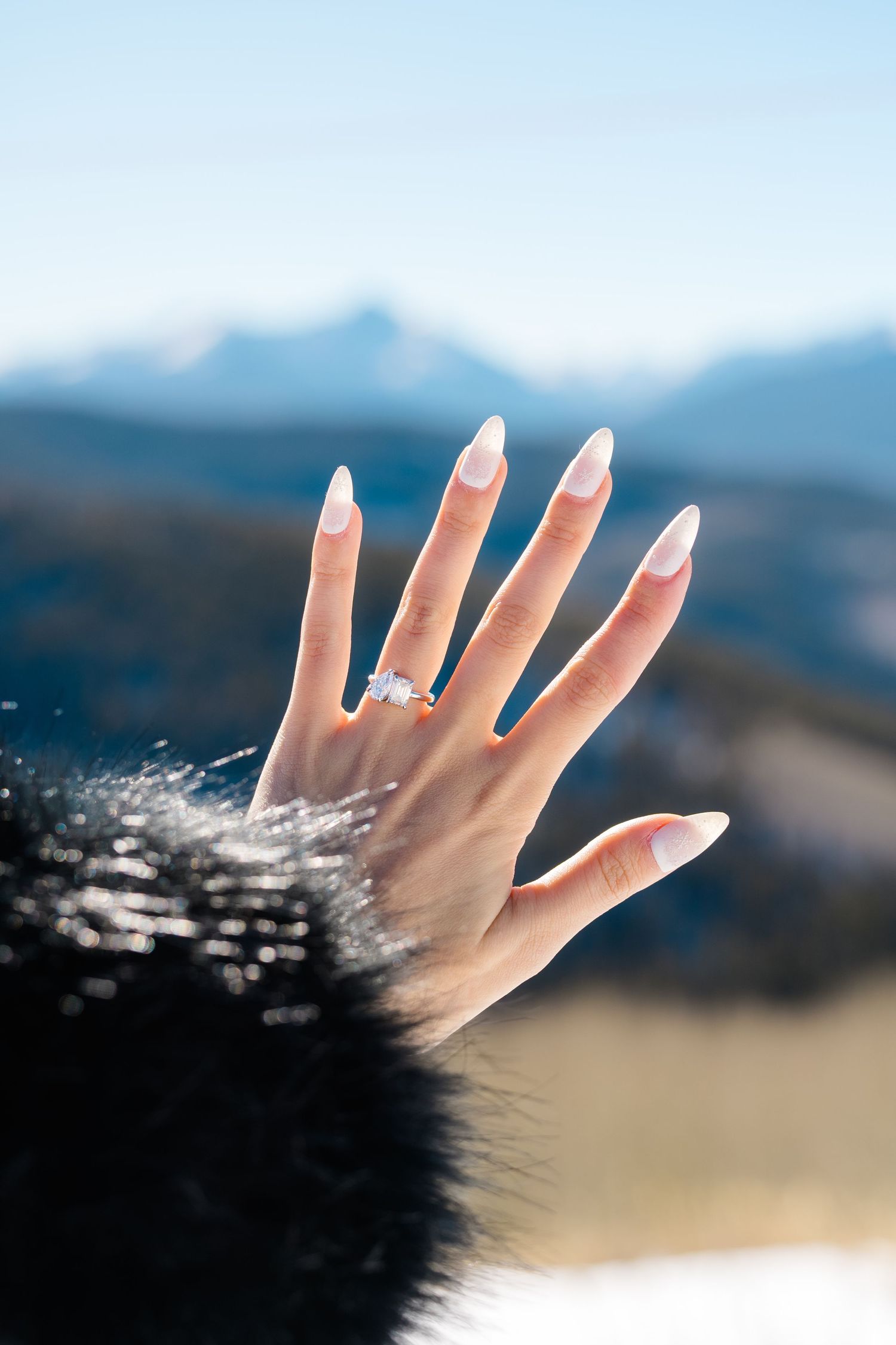 Hand with engagement ring shown against blurry mountain landscape backdrop in soft natural lighting.