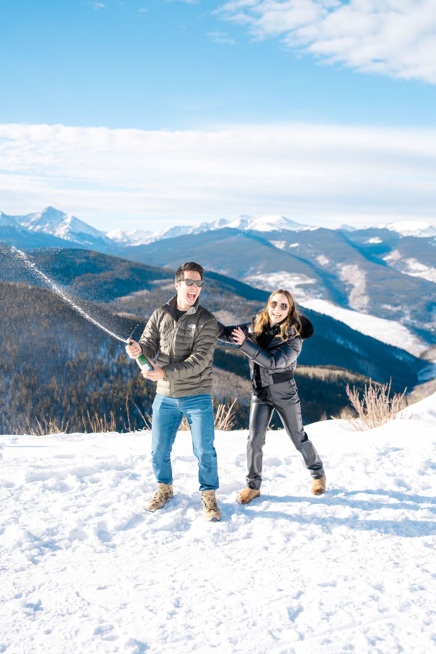 Two people pose playfully in winter attire on a snowy mountain peak with dramatic Rocky Mountain vistas in the background.