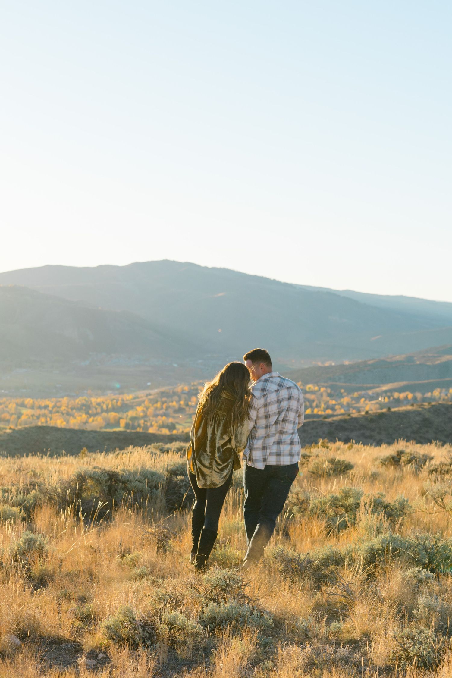 Two people stand together on a golden hillside at sunset with mountains in the background.