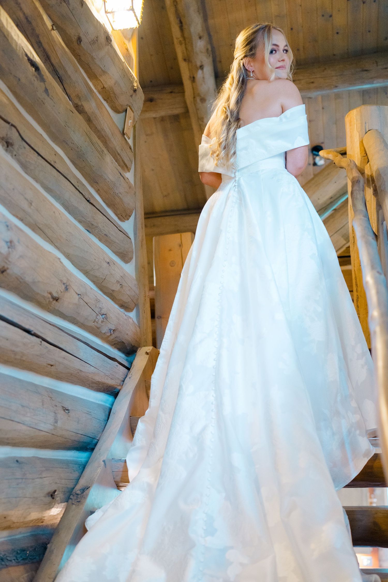 Bride in flowing white wedding gown poses in rustic wooden attic setting with natural light streaming through windows.