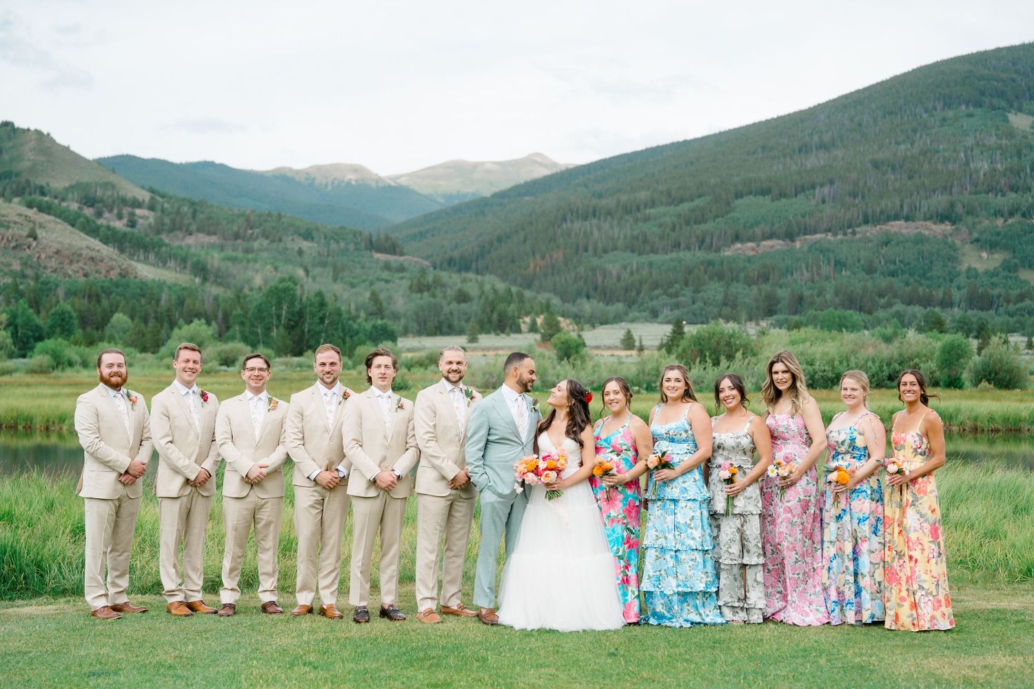 Wedding party members stand in a row against a scenic mountain backdrop with rolling green hills and distant peaks.