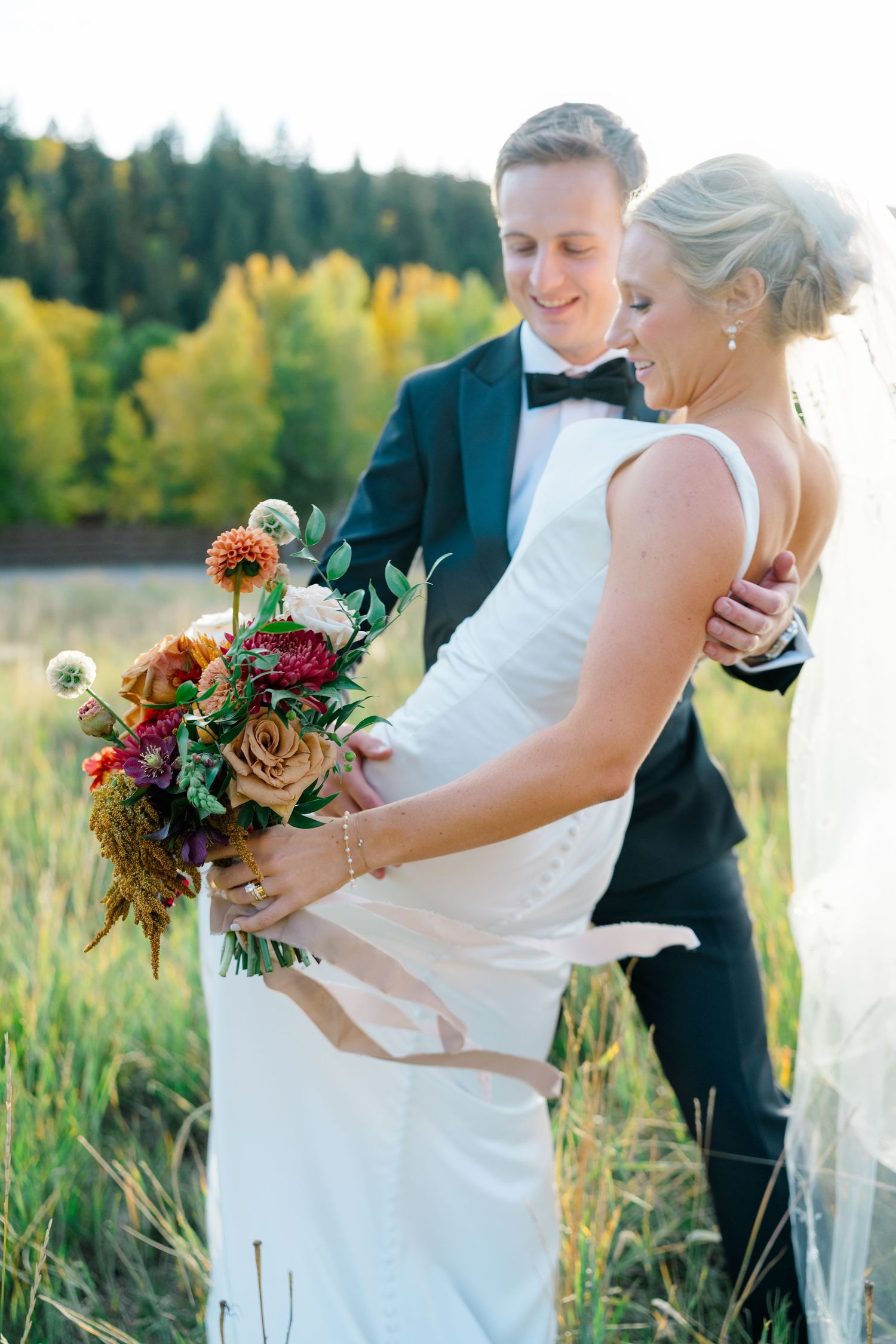 Newlyweds share romantic moments in a sunlit meadow with fall foliage and rustic bouquet.