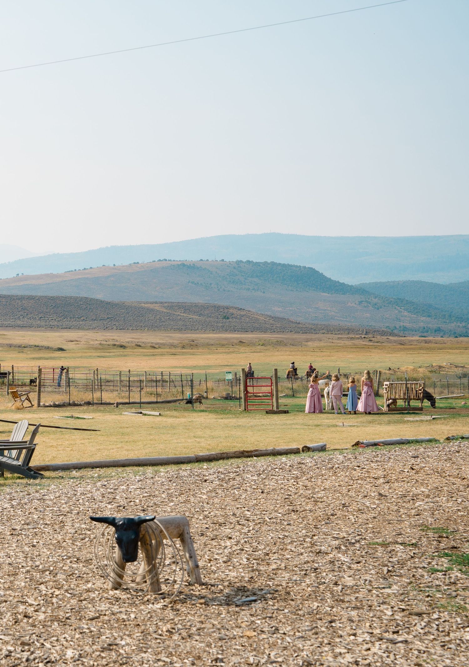 A dog stands in a dusty field with mountains and colorful figures in the background at a rural ranch setting.