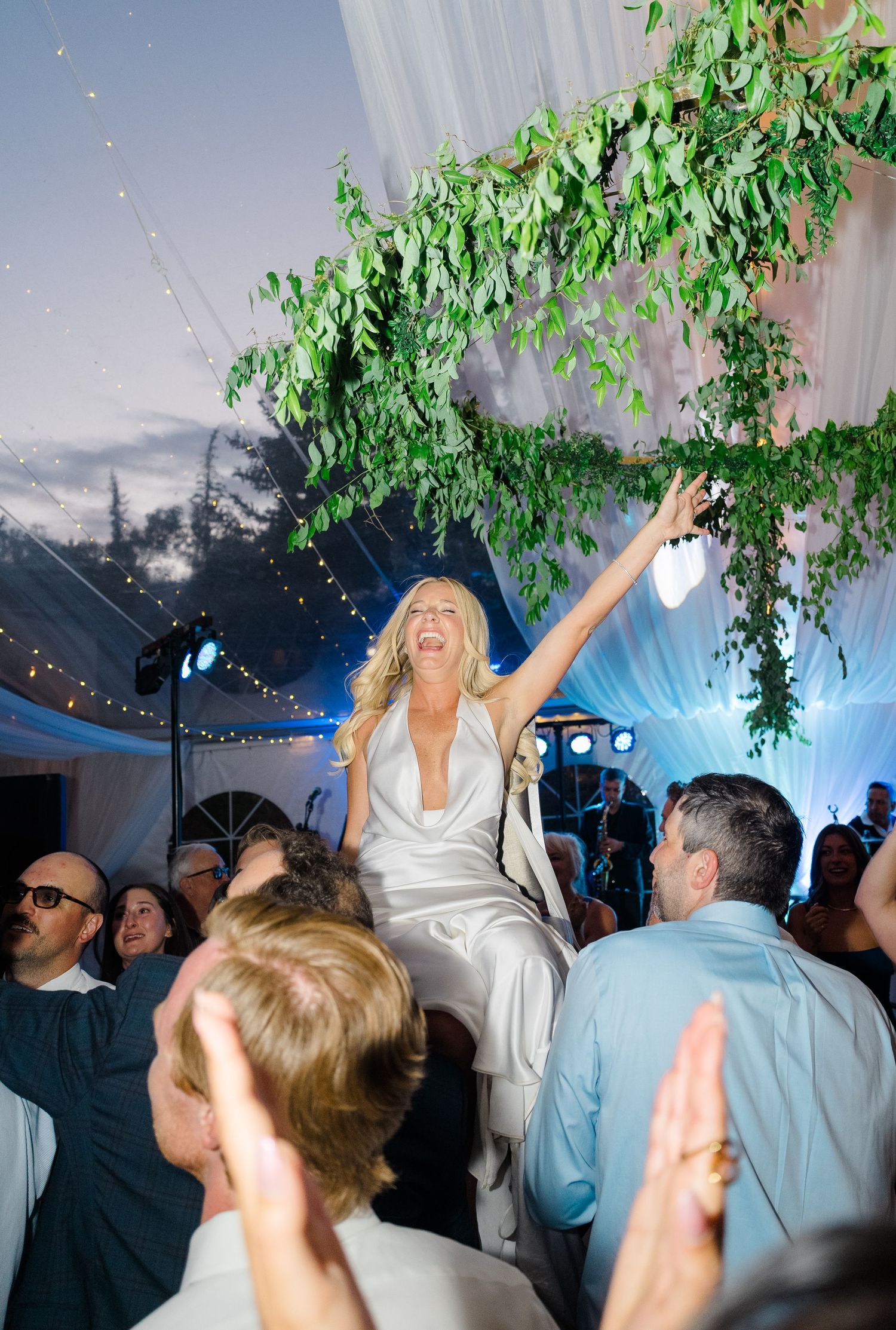 Bride in white gown raised by guests during lively wedding reception under floral greenery canopy and string lights.