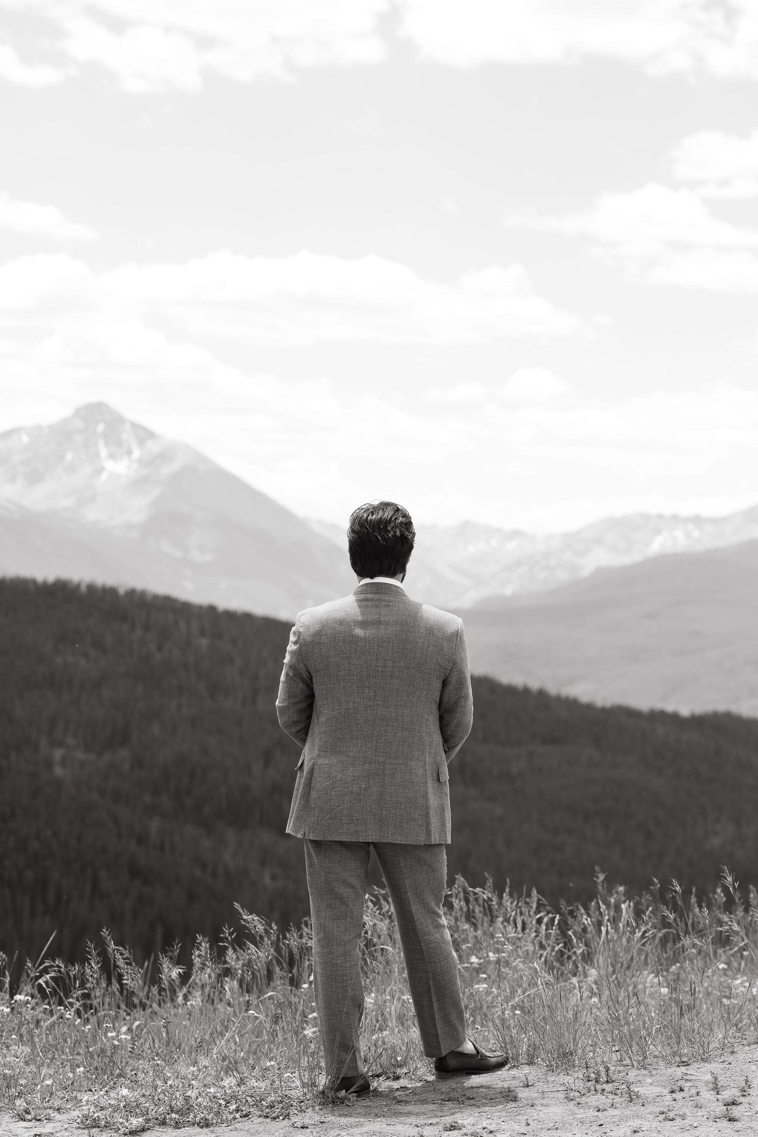 Black and white photo of a man in a suit standing on a hillside gazing at a majestic mountain landscape.