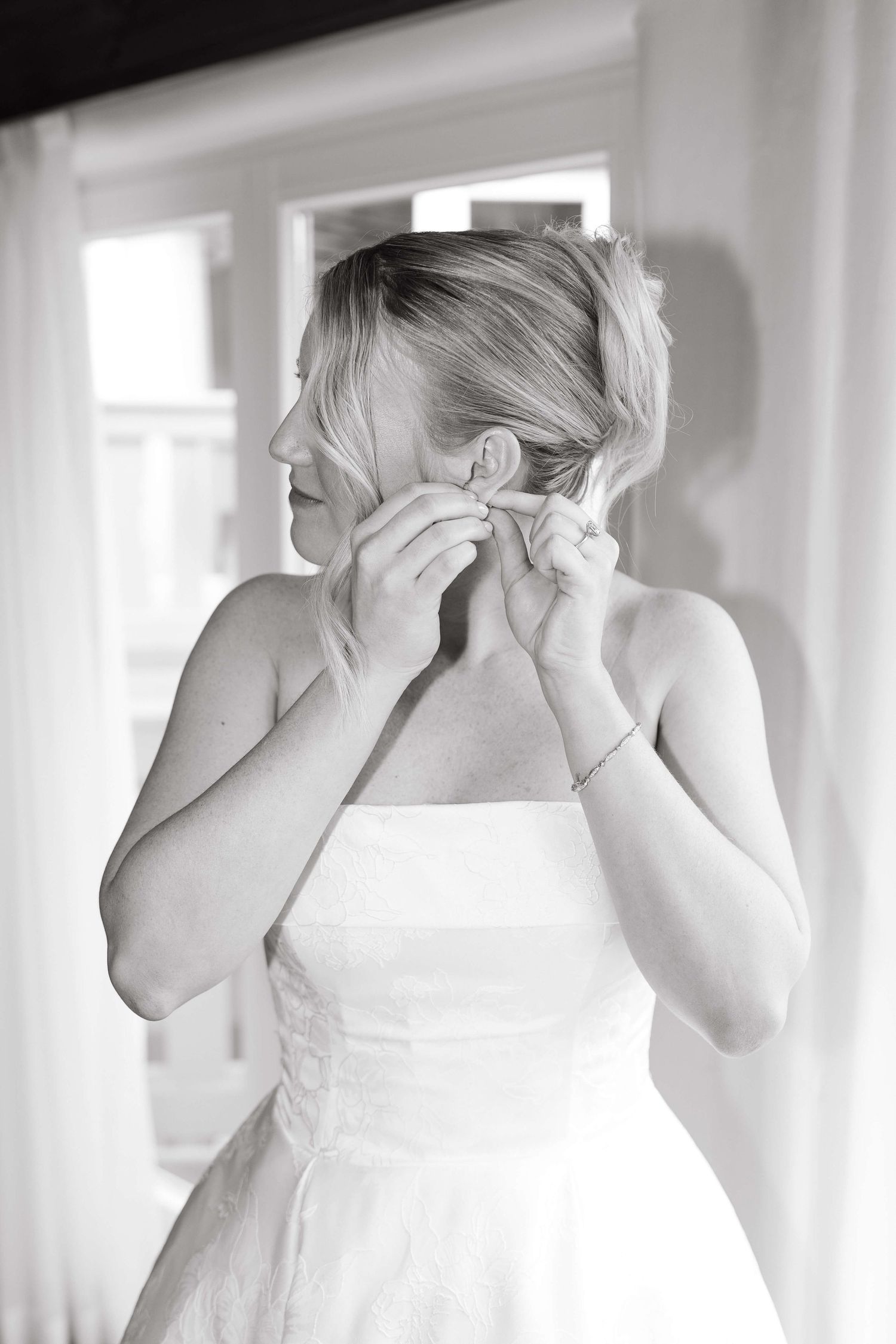 Bride putting on earrings in black and white photo wearing strapless wedding dress.