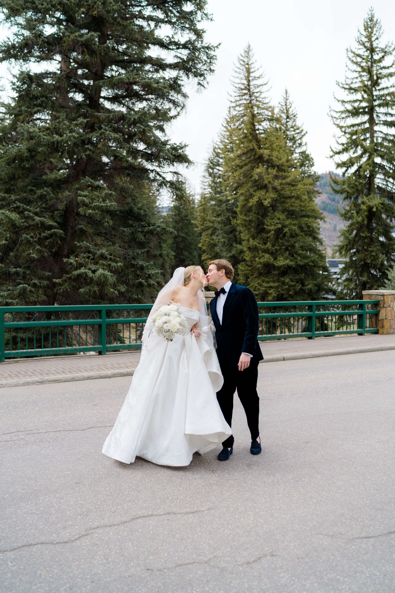 Bride and groom share a kiss on a road surrounded by tall evergreen trees in a mountain wedding setting.