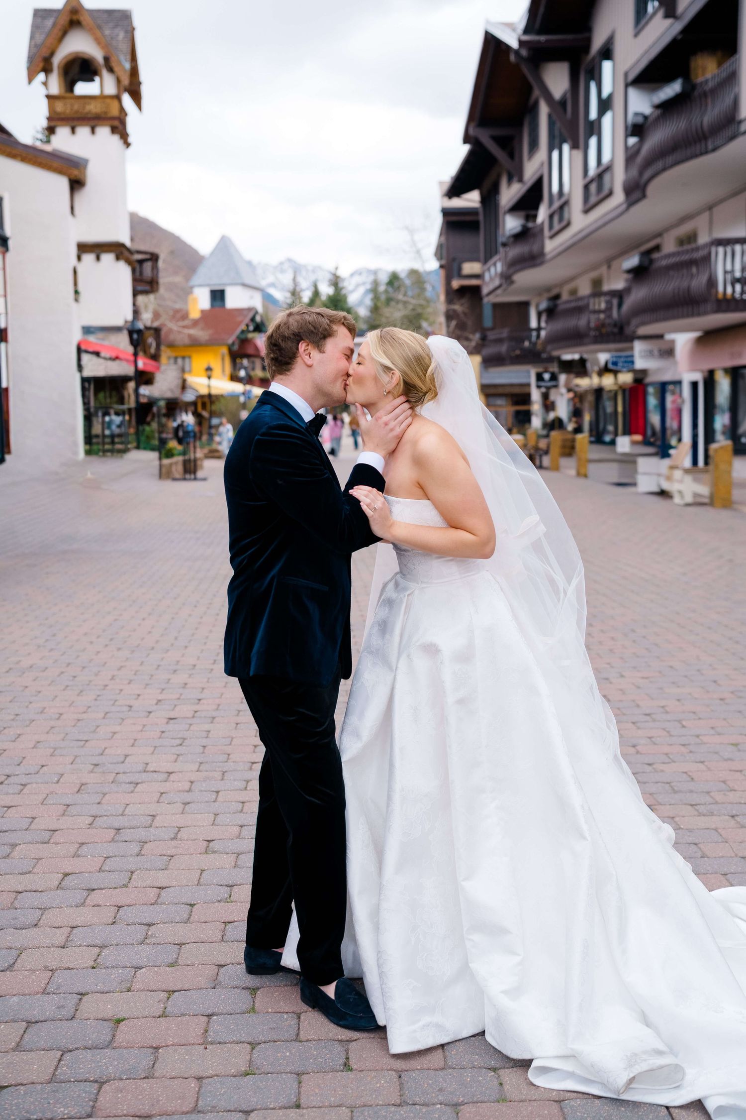 Bride and groom share a romantic kiss in a charming mountain village square, bride in white gown with veil.