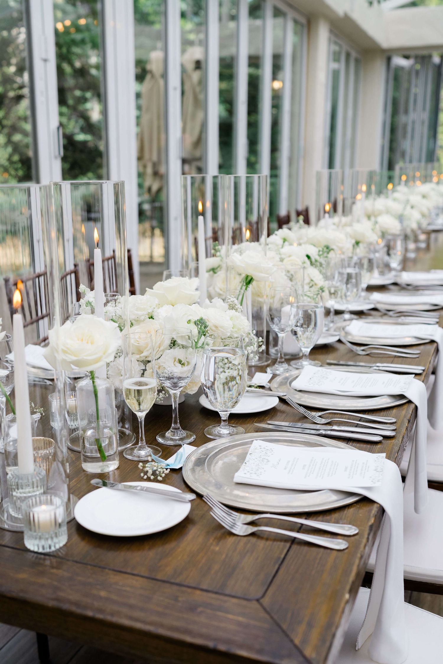 Elegant wedding reception table with white floral centerpieces, crystal glassware, and silver charger plates on rustic wood.