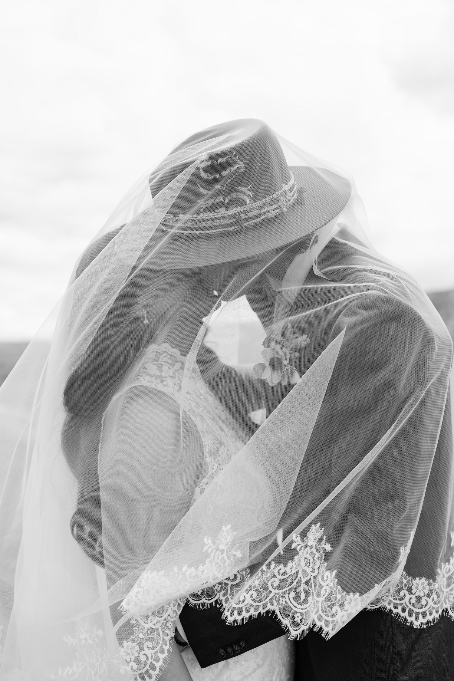 Black and white photo of bride and groom kissing under lace wedding veil, groom wearing stylish hat.