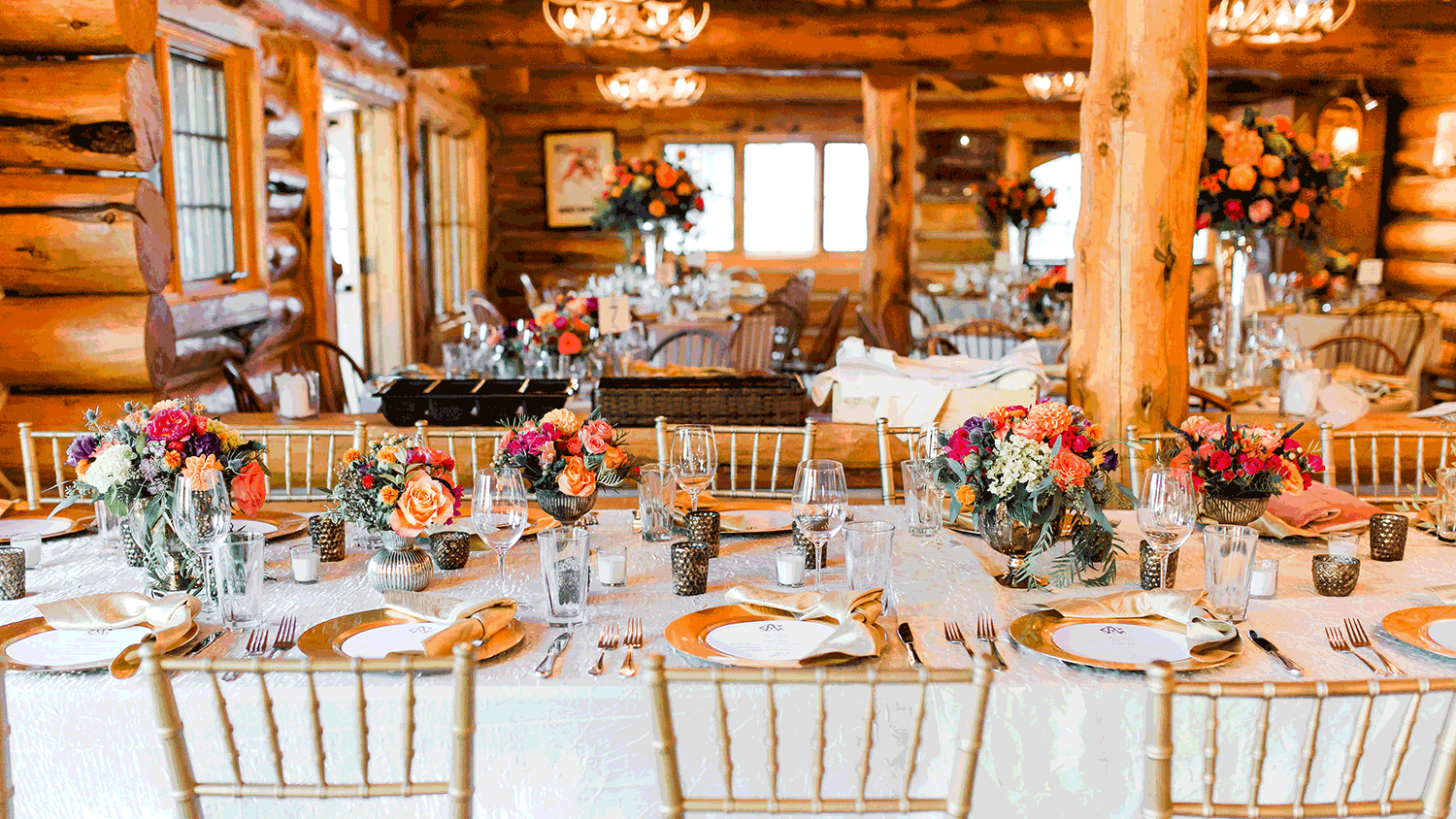 Elegant rustic log cabin wedding reception with gold chiavari chairs, white linens, and vibrant floral centerpieces.