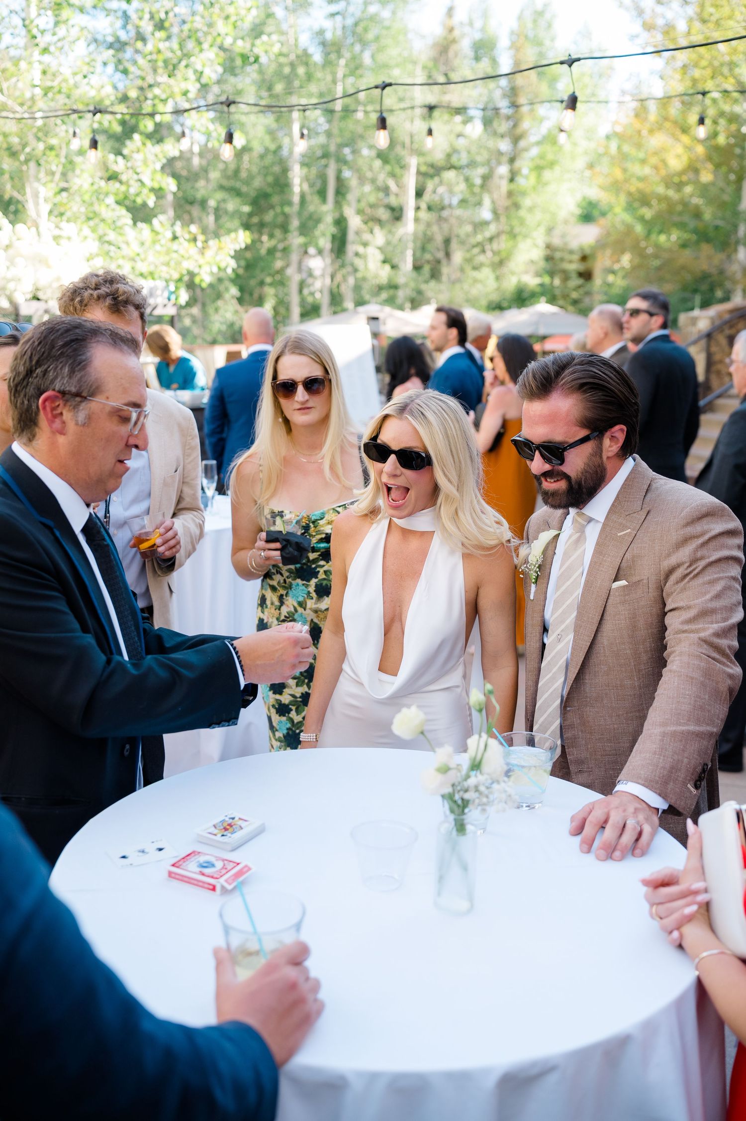 Guests react with amazement during a card magic trick at an elegant outdoor wedding cocktail hour.