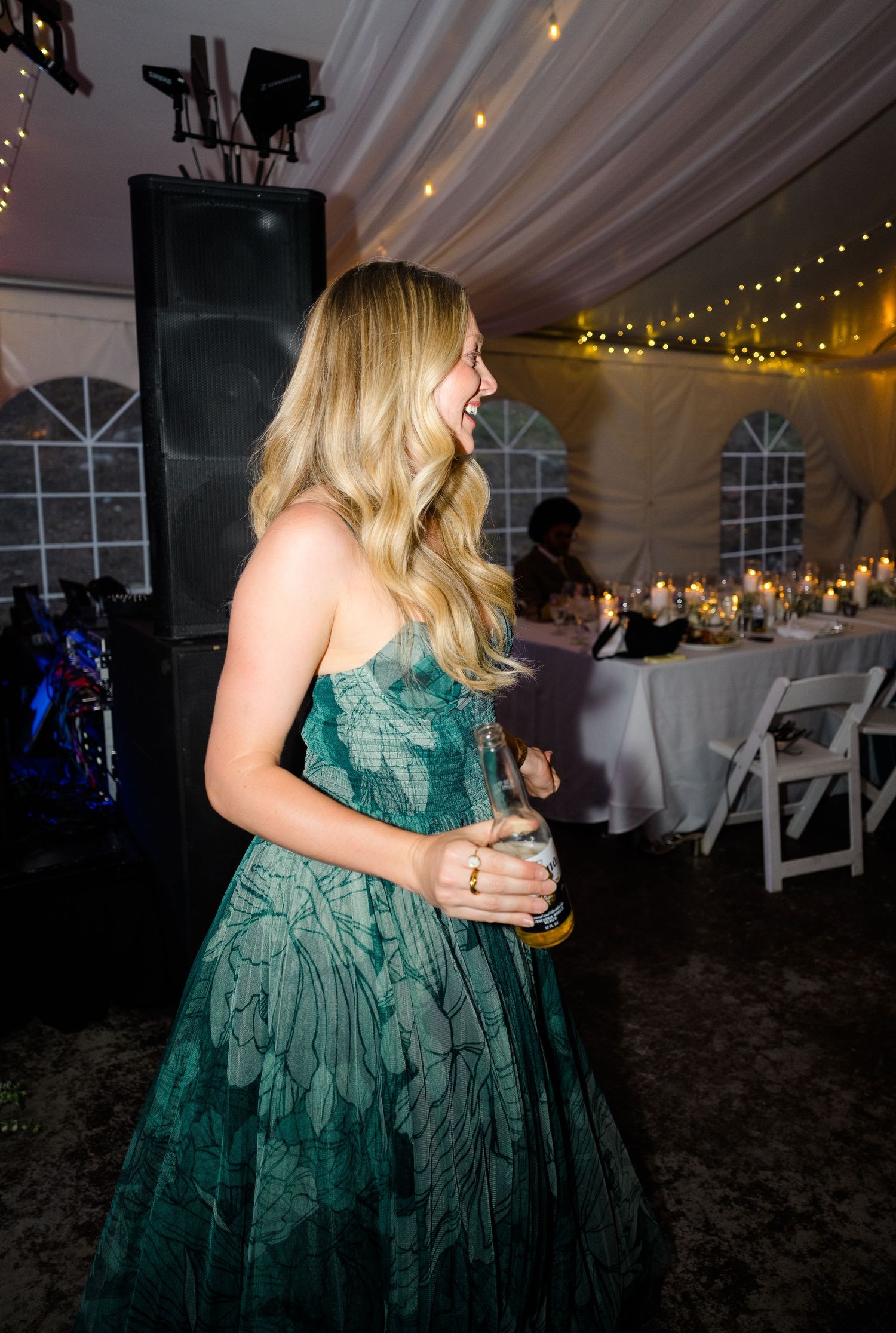 A woman in a strapless green floral gown laughs while holding a drink at an elegant tented wedding reception.
