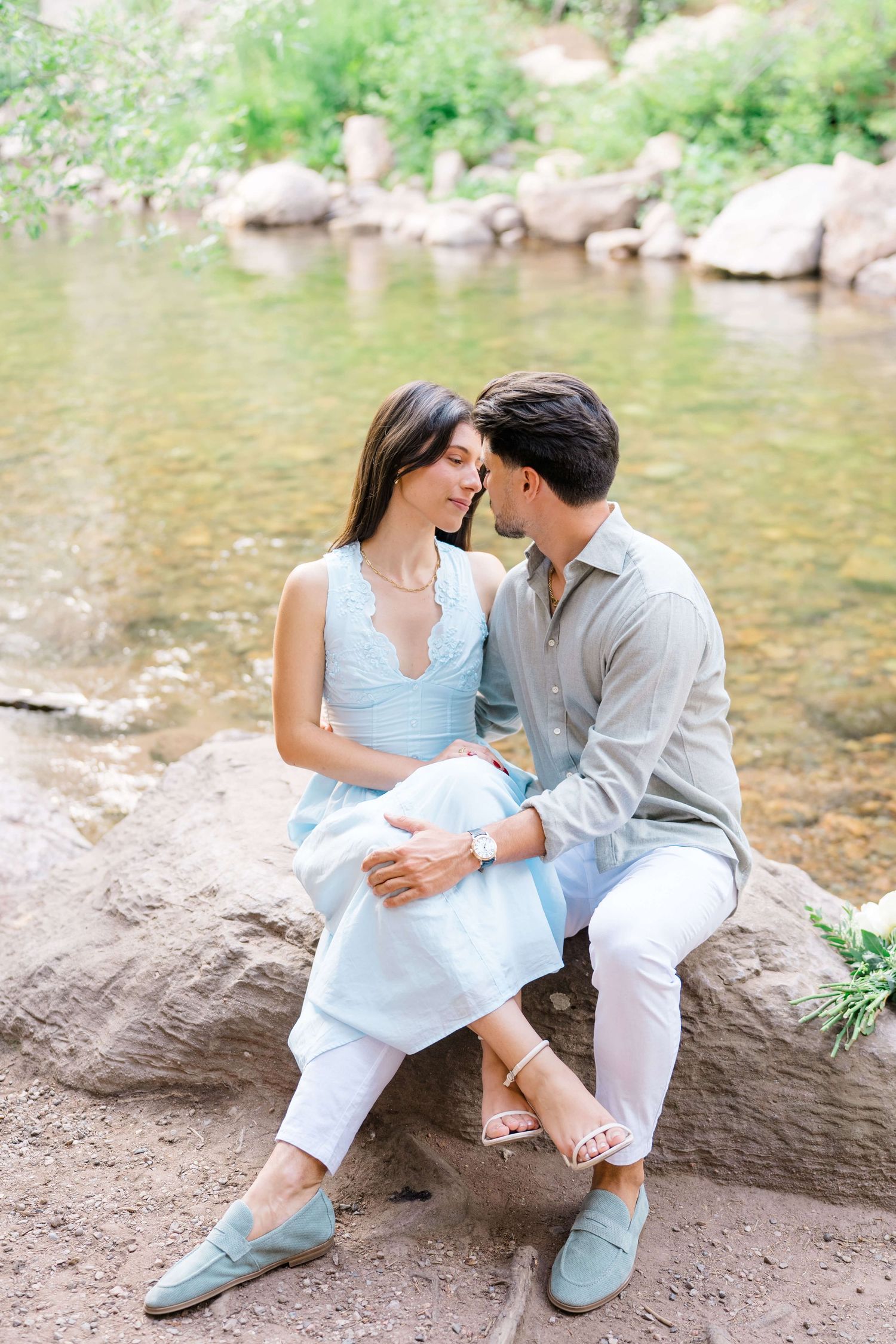 After a vail village proposal, a couple in light blue and white outfits sit intimately together on rocks by a peaceful stream.