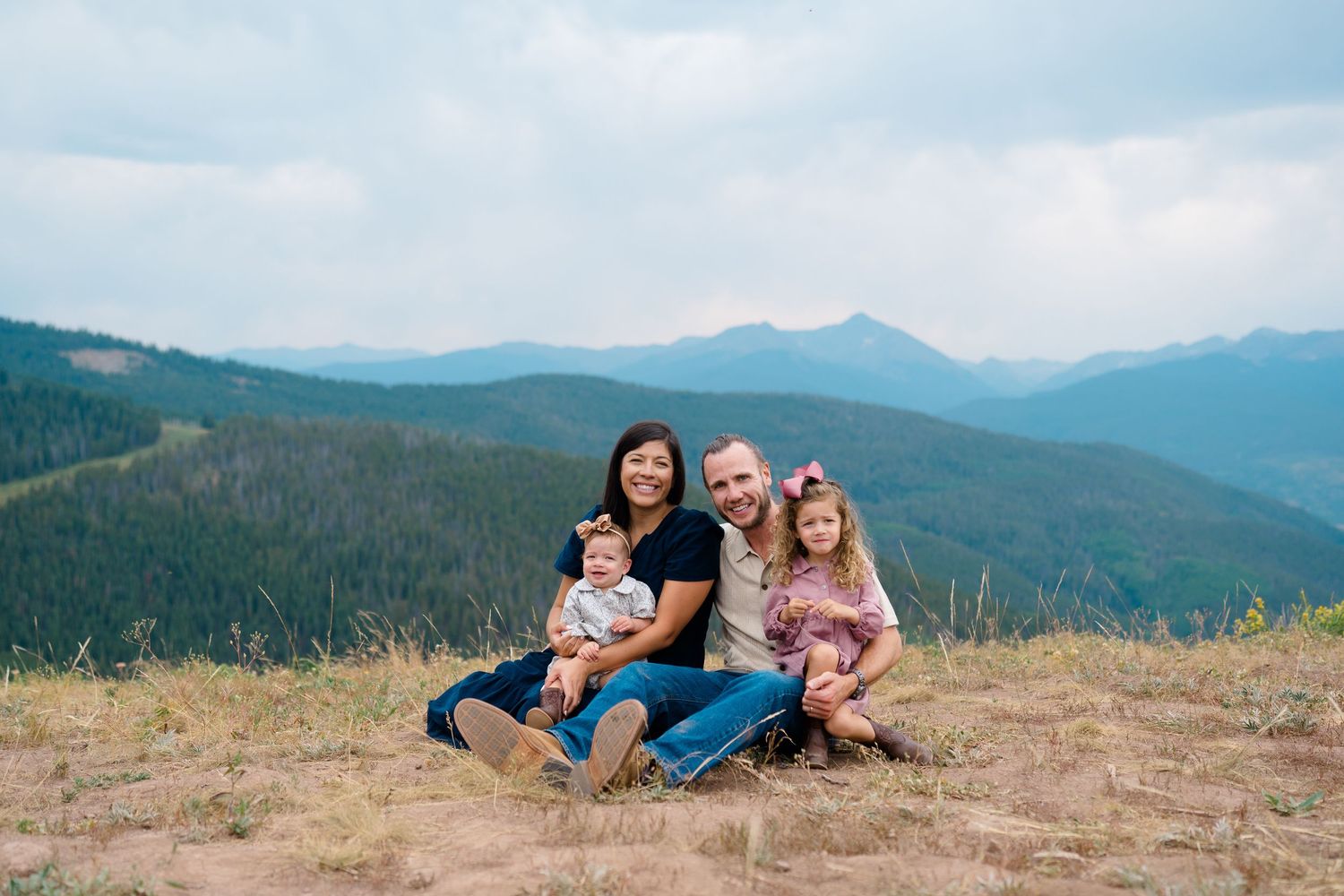 A family sits together on a grassy hillside overlooking scenic mountain vistas on a cloudy day during family mountaintop photography session