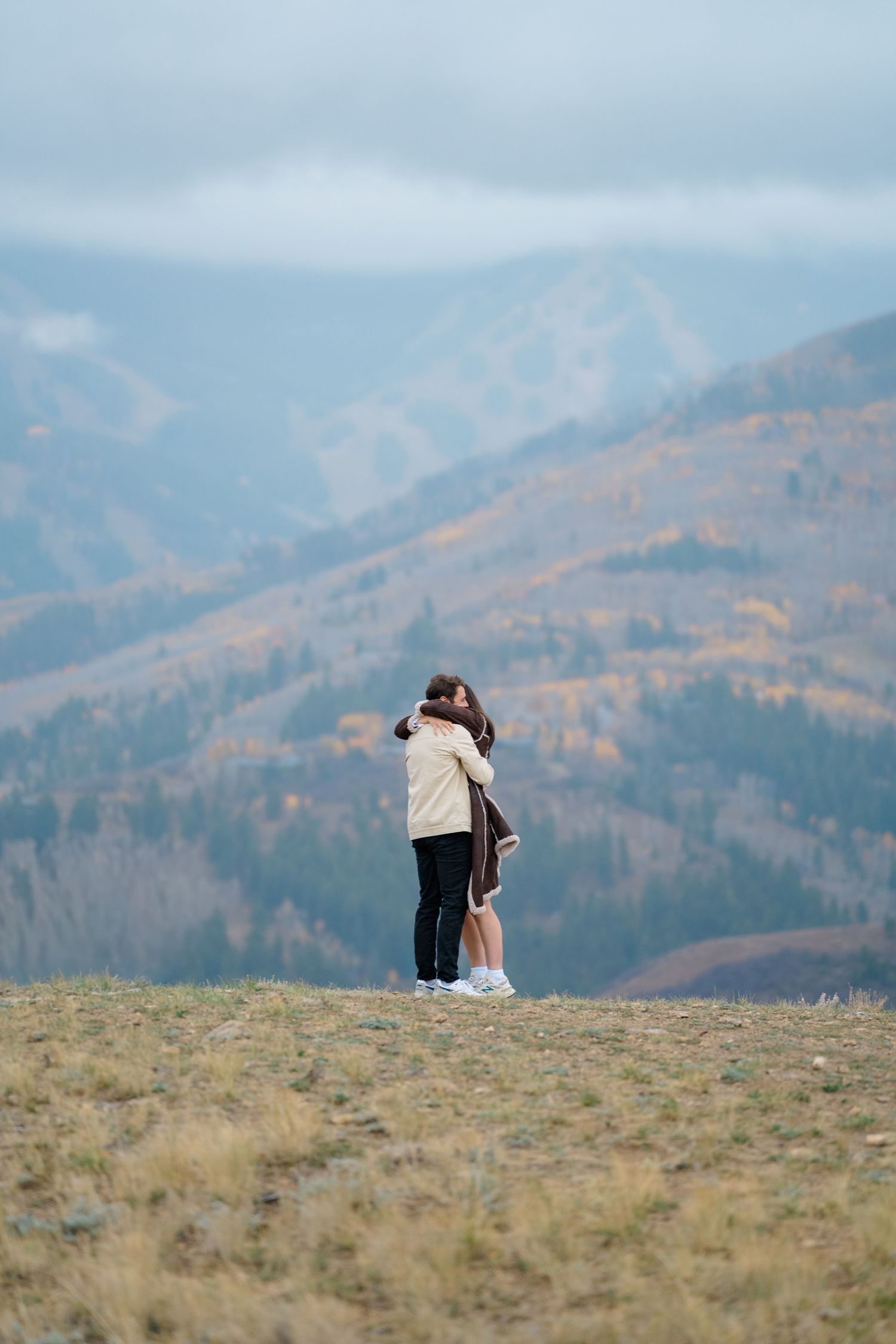 Two people embrace on a hilltop overlooking scenic mountain vistas with autumn colors in the distance.