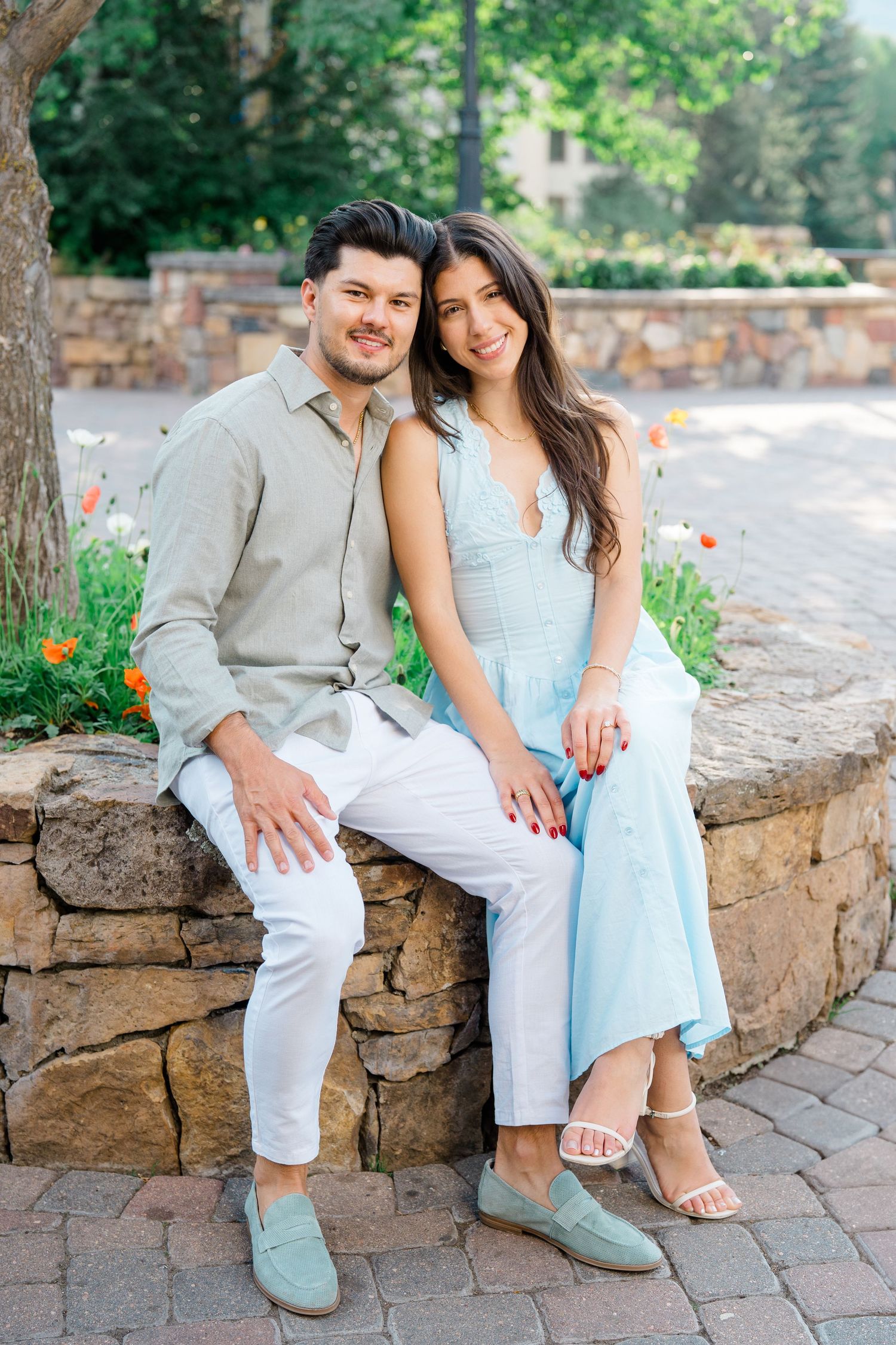 A couple sitting on a stone wall in coordinating light blue and white outfits in a garden setting.
