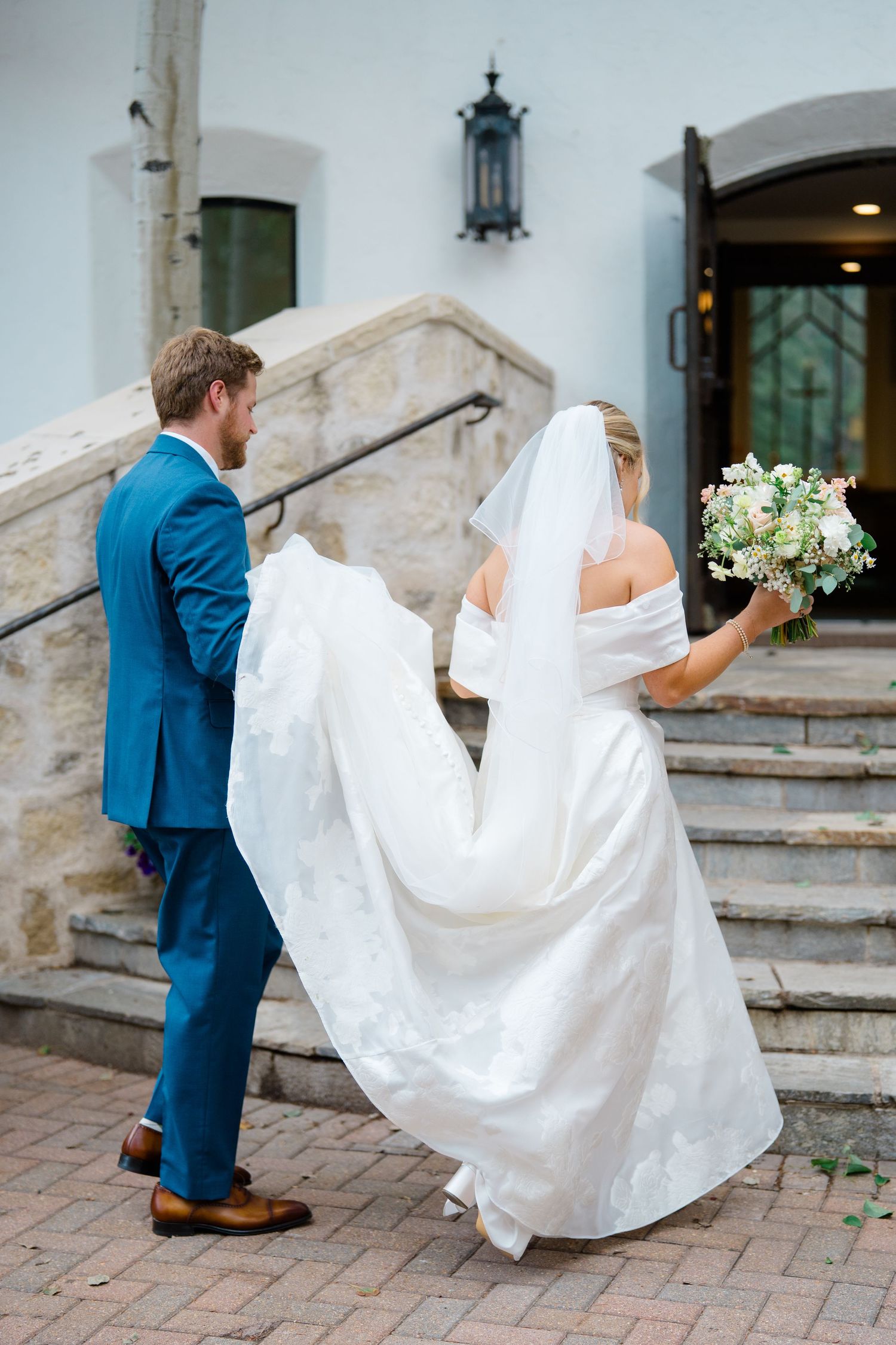 A couple in wedding attire walks up stone steps with the bride's white dress flowing elegantly behind them during vail wedding prep.