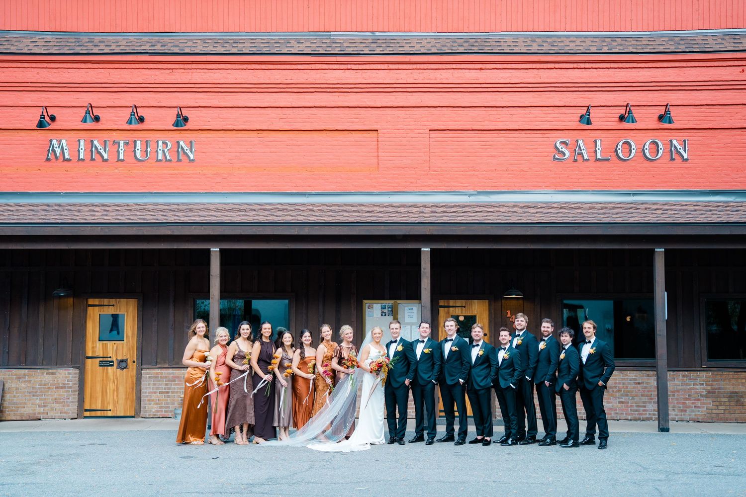 Wedding party lines up outside red-trimmed building for formal group photo.