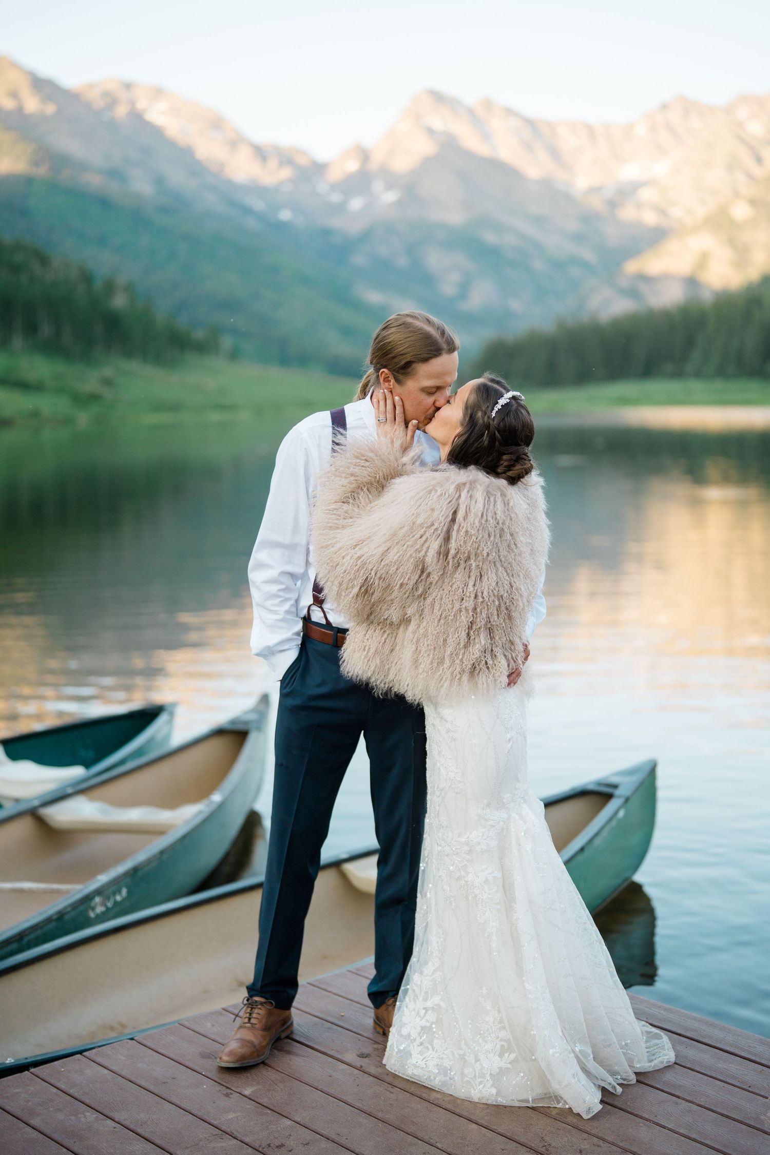 Wedding couple embracing on a wooden dock with mountain lake and canoes in the background at sunset.