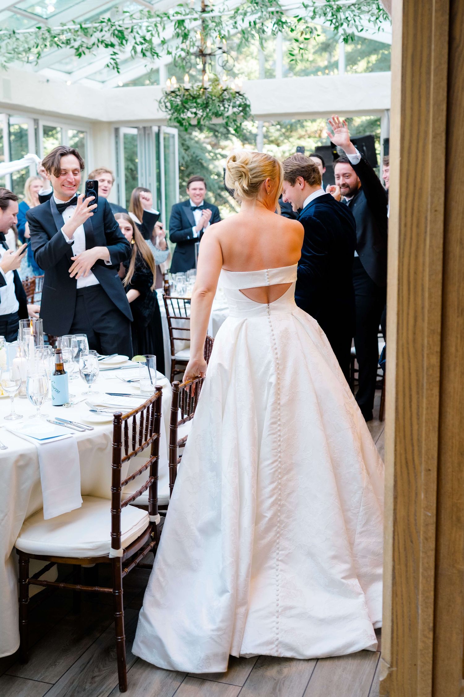 Bride in strapless ball gown enters wedding reception as guests cheer in elegant greenhouse venue.