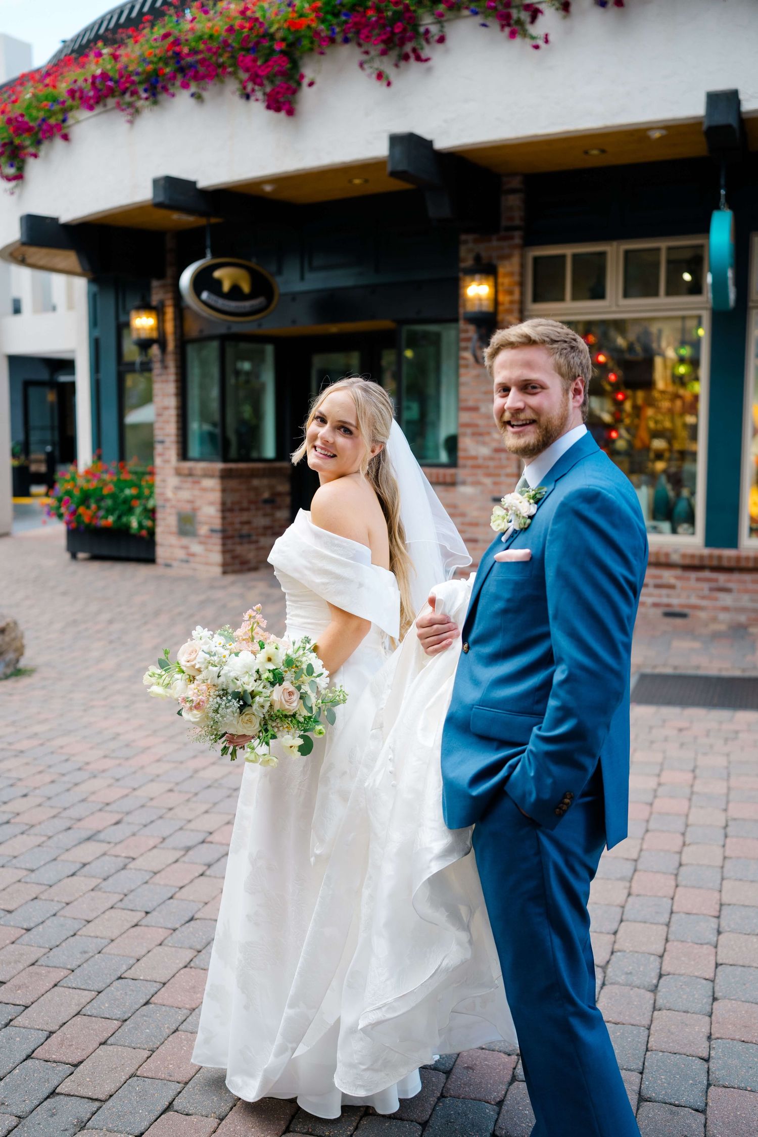 Bride and groom walk along cobblestone path with hanging flower baskets and storefronts at vail wedding.