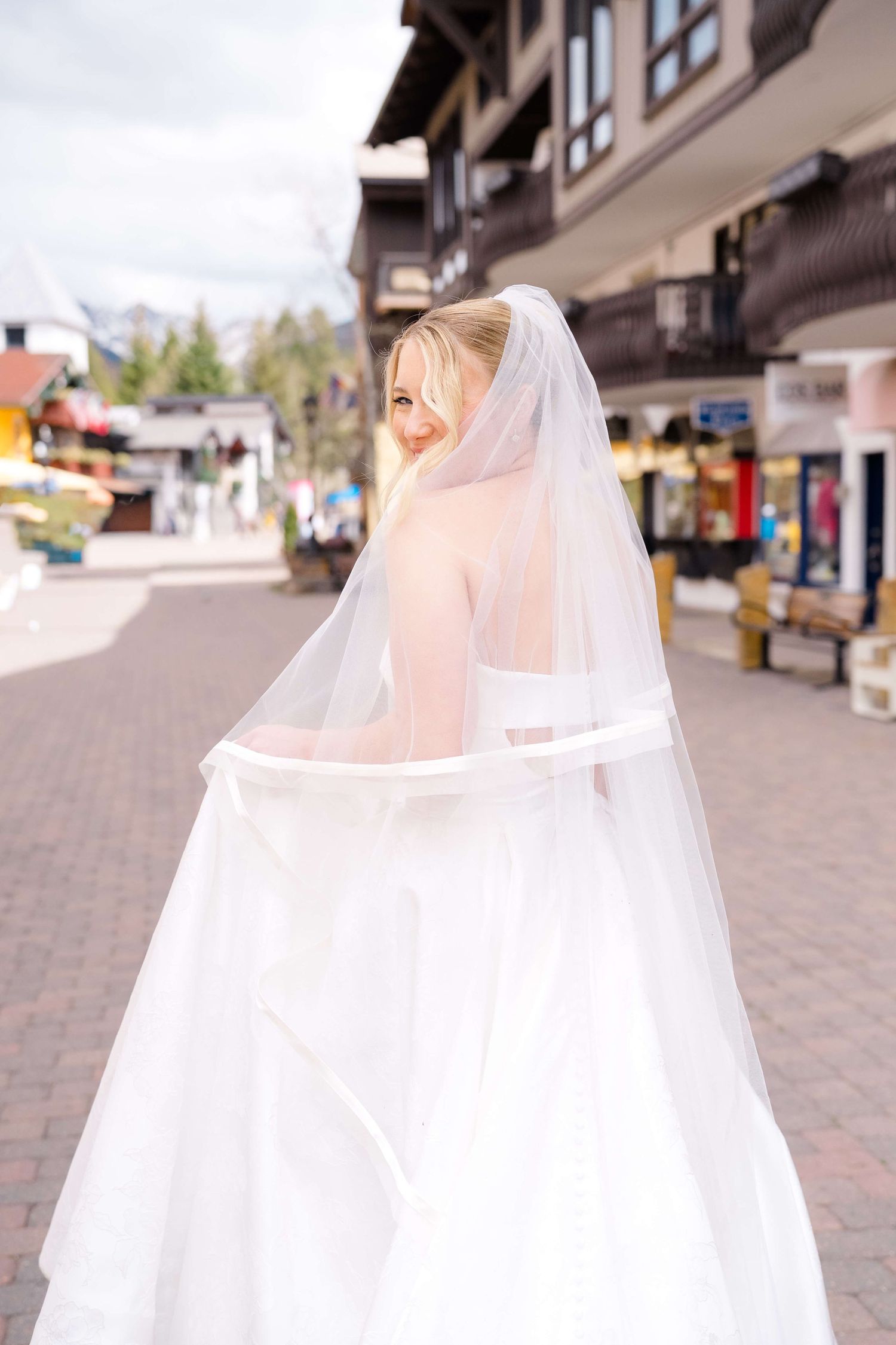 Blonde bride in white gown and veil walking through outdoor shopping street, looking back over shoulder.