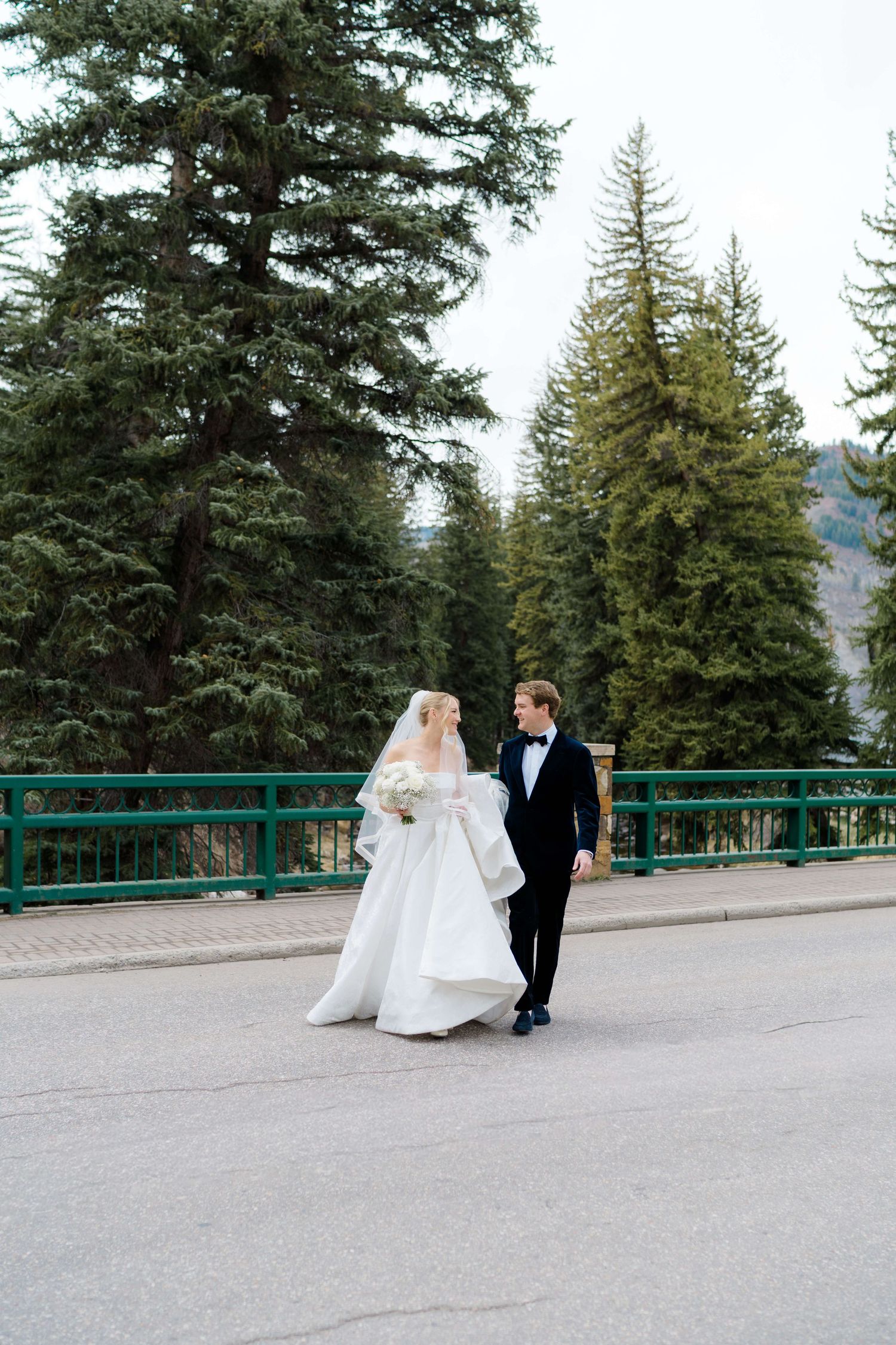 Bride and groom walk hand-in-hand on a bridge surrounded by tall evergreen trees in a mountain setting.